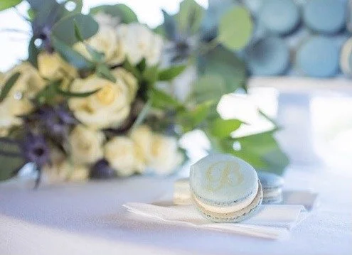 A white macaron with light green filling on a white napkin, with a bouquet of white and purple flowers in the background.