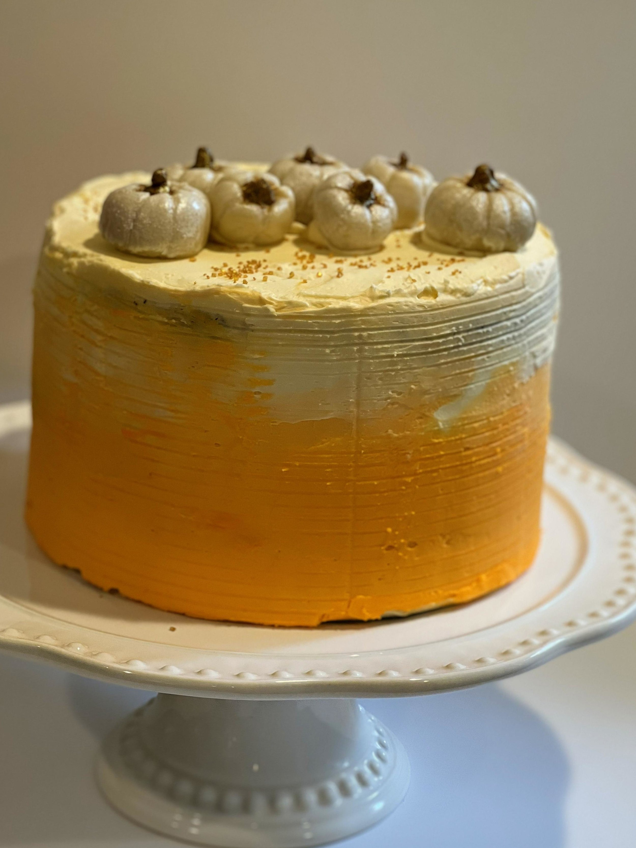 A layered cake with white frosting and decorated with white pumpkin-shaped decorations on top, placed on a white cake stand.