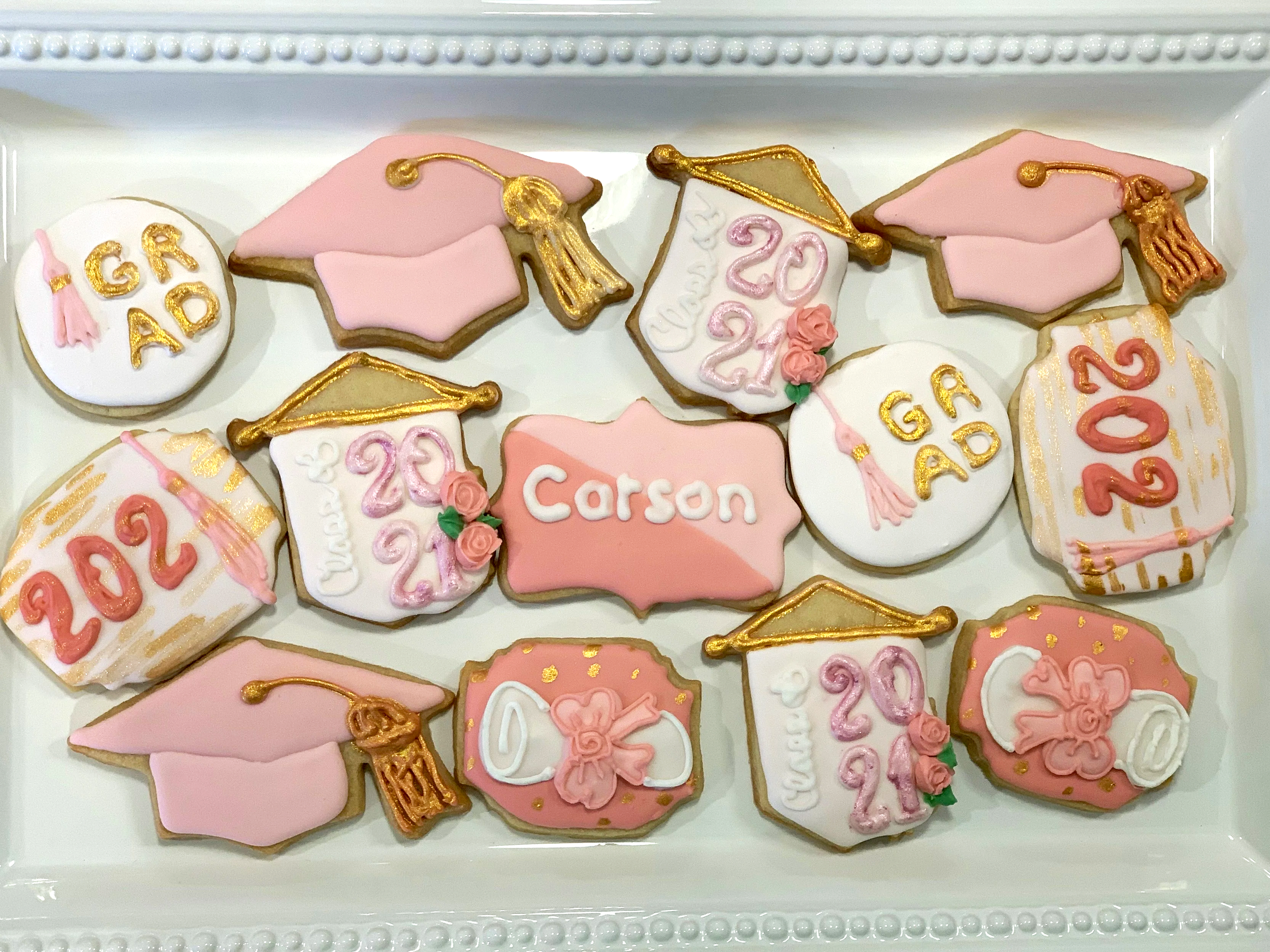 Collection of decorated graduation cookies, including diplomas, graduation caps, and certificates, with pink, white, and gold accents.