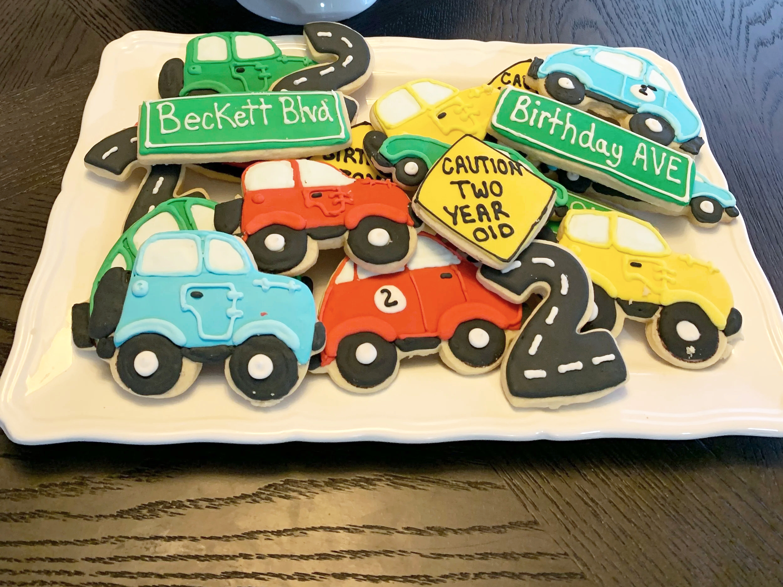 Decorated cookies shaped like colorful cars, traffic signs, and road markings on a white platter for a birthday celebration.