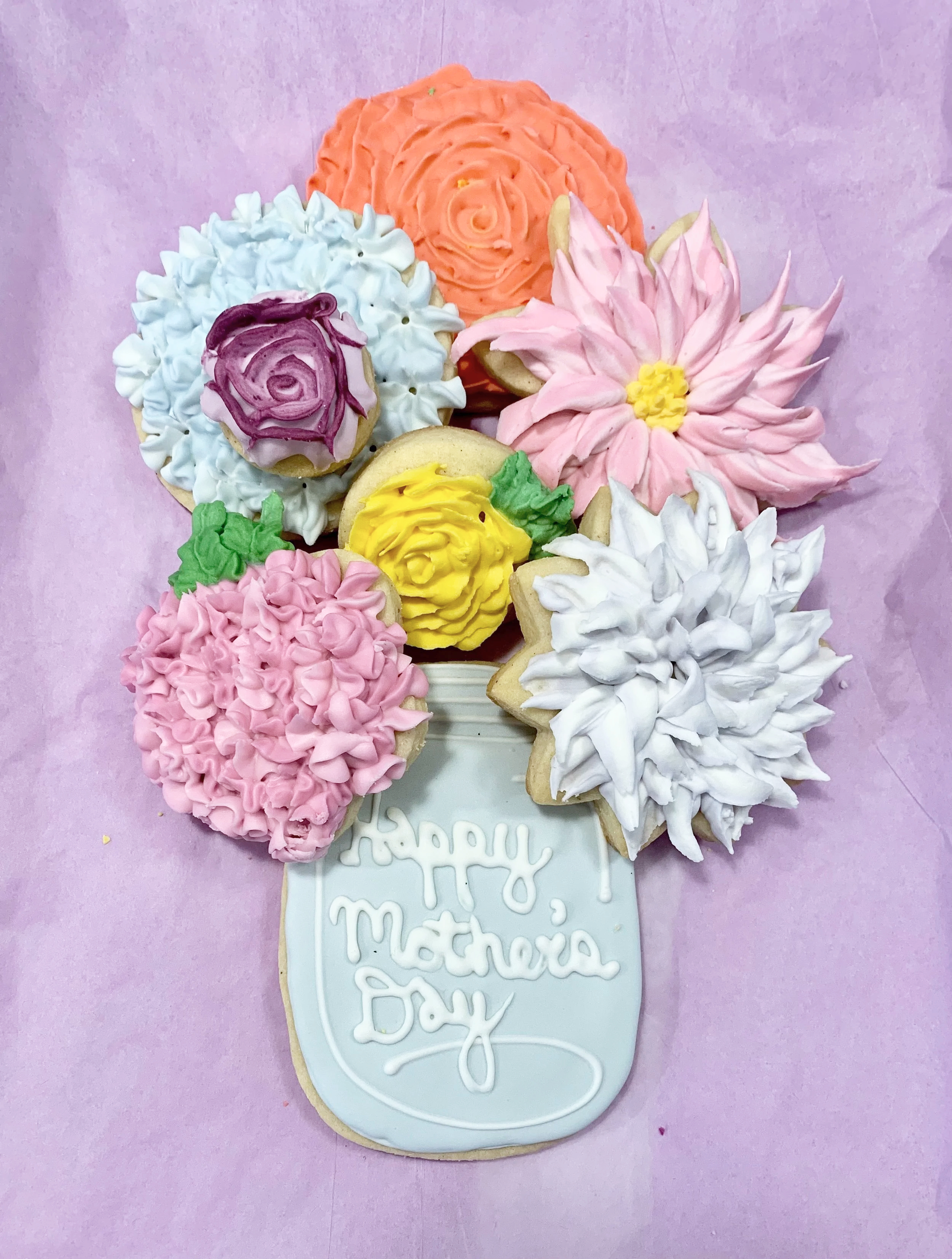 Assorted decorated cookies arranged in a bouquet shape with a cookie at the bottom that reads 'Happy Mother's Day' on a light pink background.
