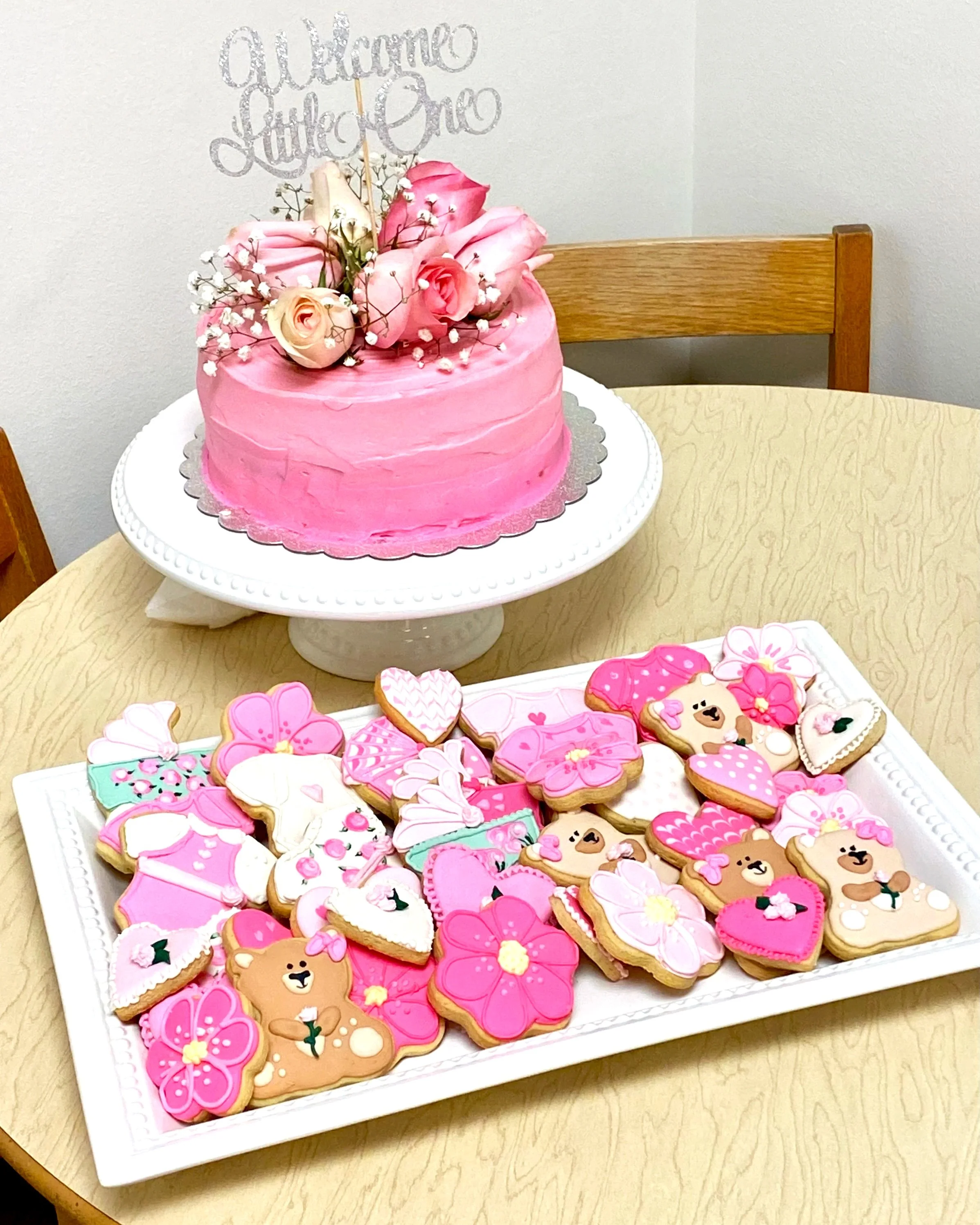 Pink frosted cake decorated with flowers and a silver 'Welcome Little One' topper, placed on a white cake stand. In front, a tray of decorated cookies with pink, white, and brown icing, featuring bows, flowers, teddy bears, and hearts, on a wooden ta