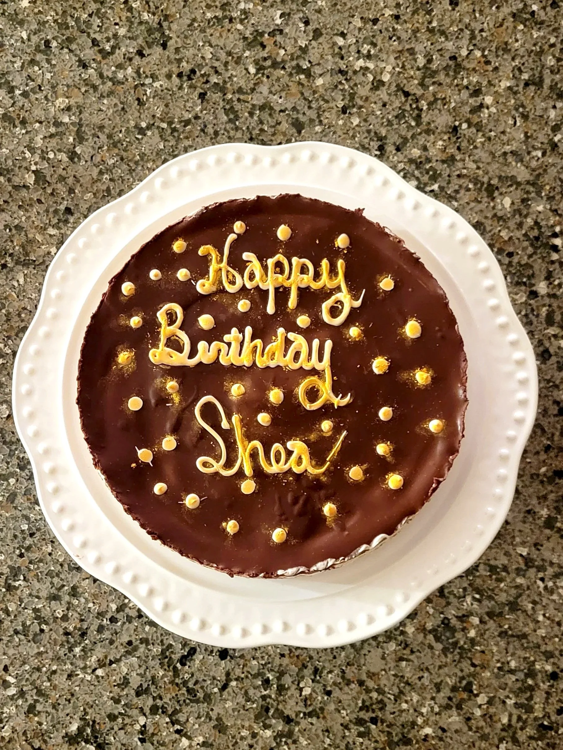 Chocolate birthday cake with yellow writing that says "Happy Birthday Shea" and decorated with small yellow dots on a white plate.
