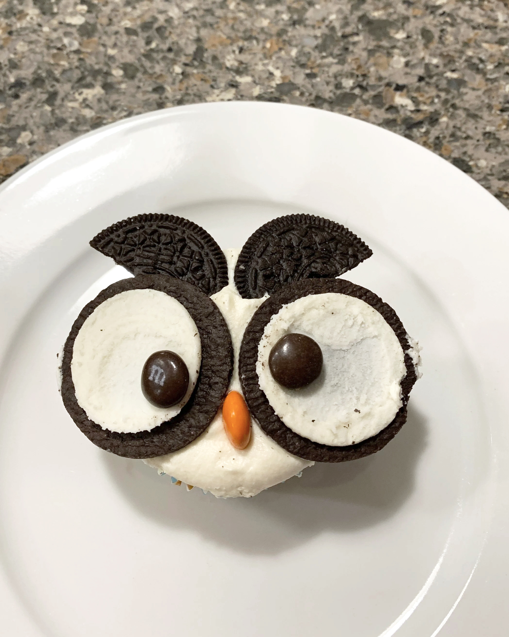 Cupcake decorated to resemble an owl, with Oreo cookies for eyes, Oreo halves for the eyebrows, a candy piece for the beak, and Oreo cookies for the ears, on a white plate.