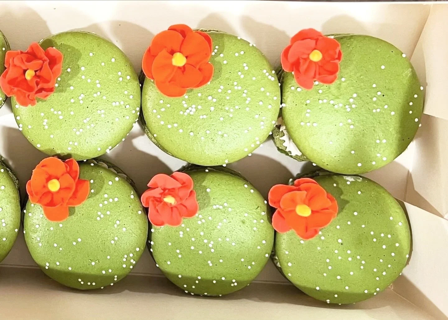 Green cupcakes decorated with small white sprinkles and orange fondant flowers with yellow centers, arranged in a box.