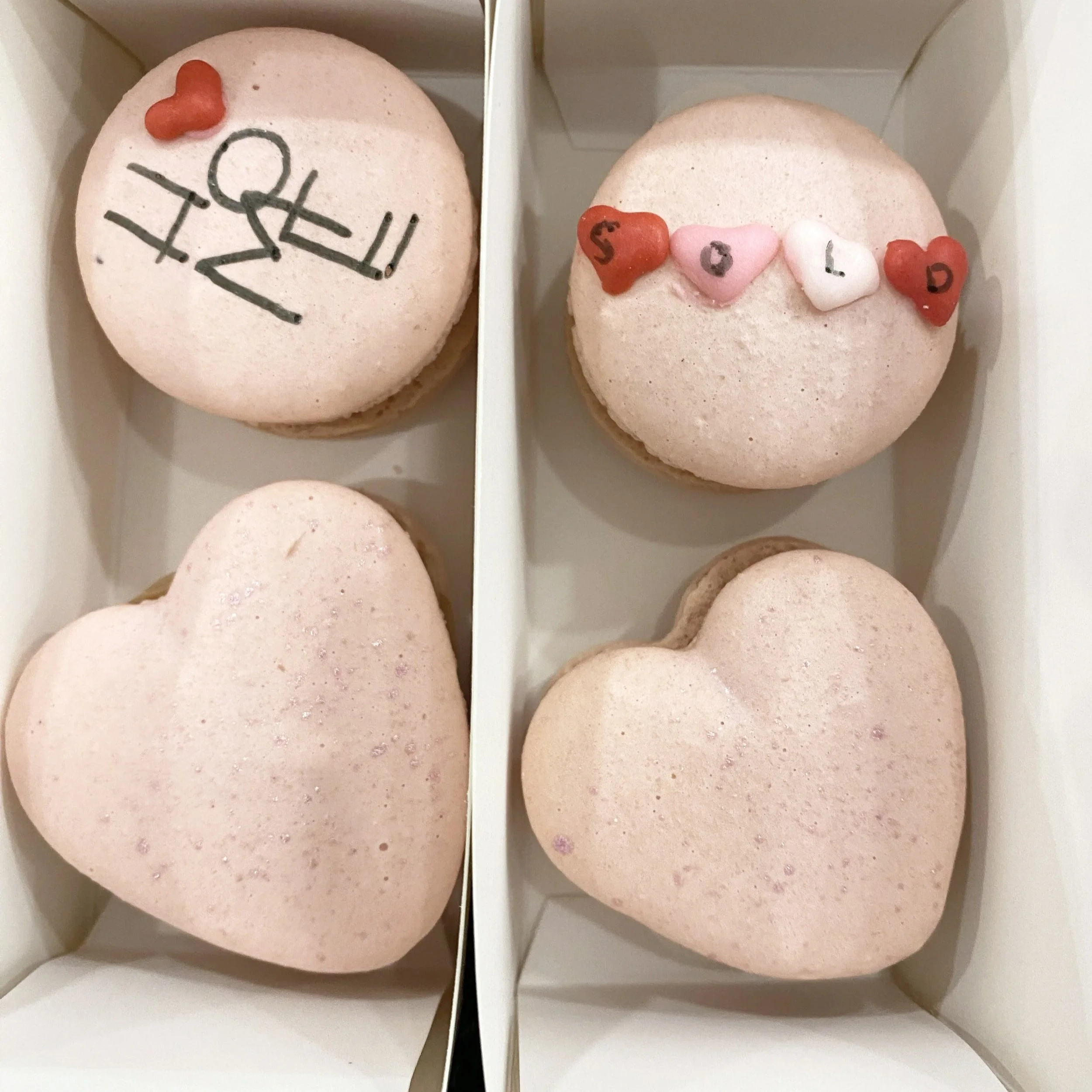 Four pink heart-shaped macarons inside a white box, with two on each side separated by a vertical divider. The top left macaron has black writing and a small red heart-shaped decoration. The top right macaron has small heart-shaped candies with the l
