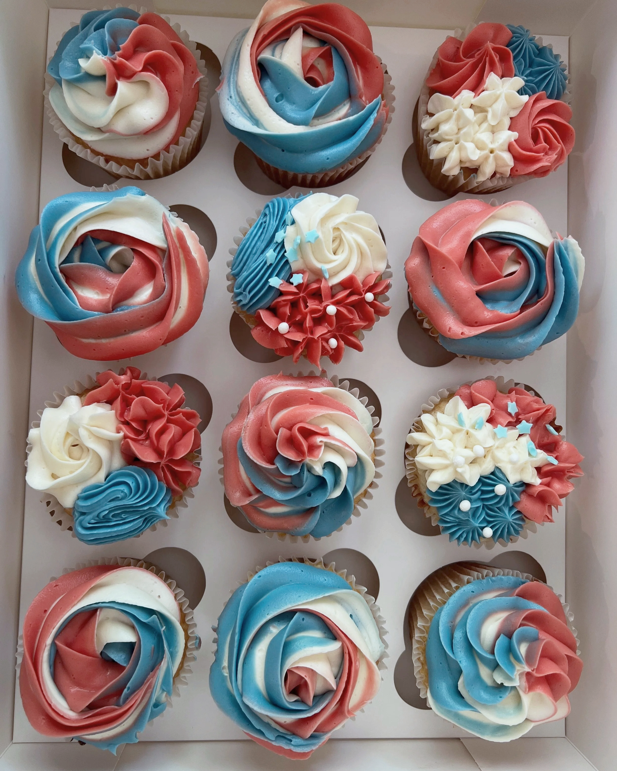 A box of twelve cupcakes decorated with red, white, and blue swirled frosting, some with additional decorative frosting flowers and stars.