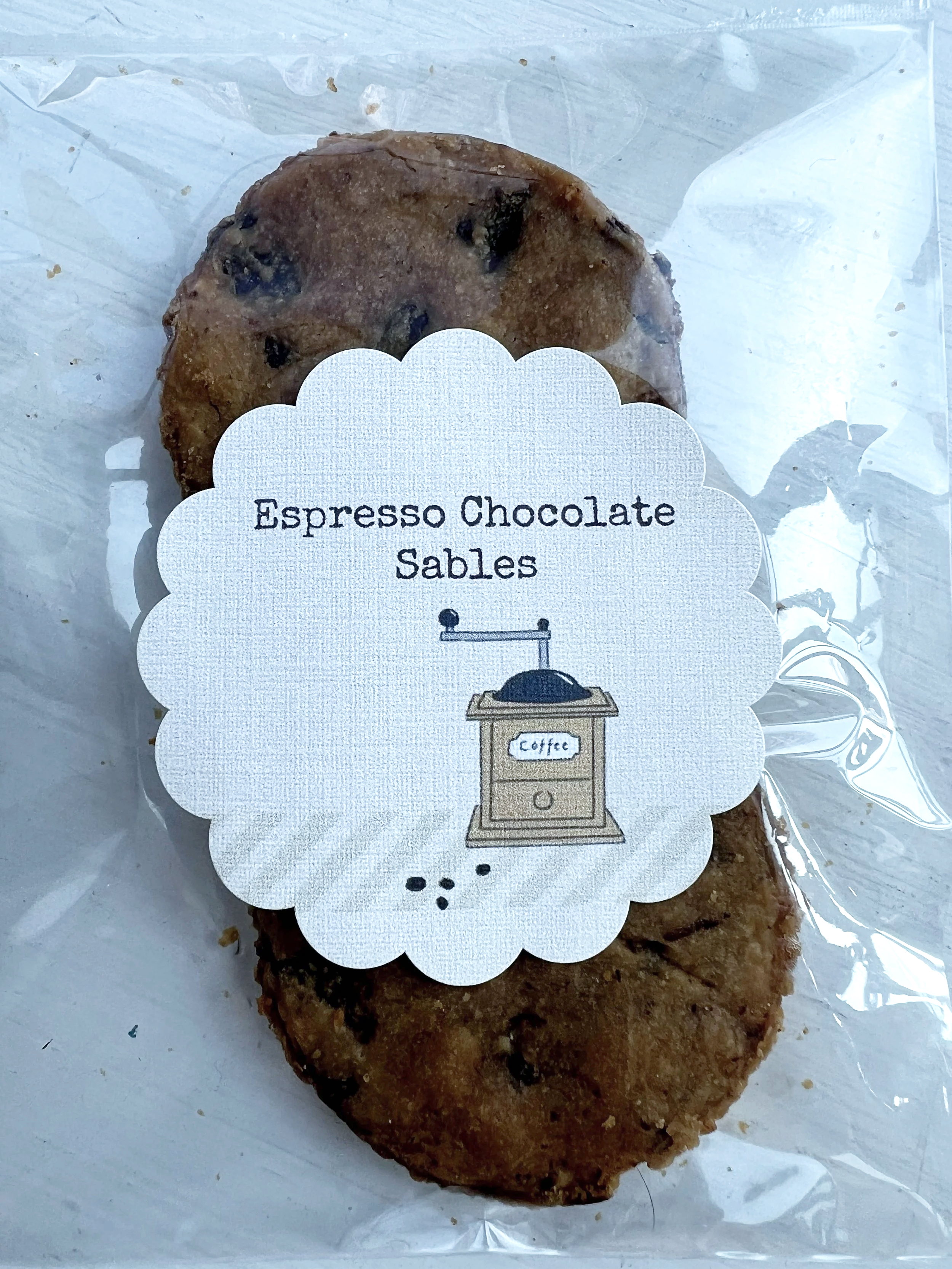 A cookie with chocolate chips wrapped in clear plastic, labeled 'Espresso Chocolate Sables' with a decorative tag featuring a coffee grinder illustration.