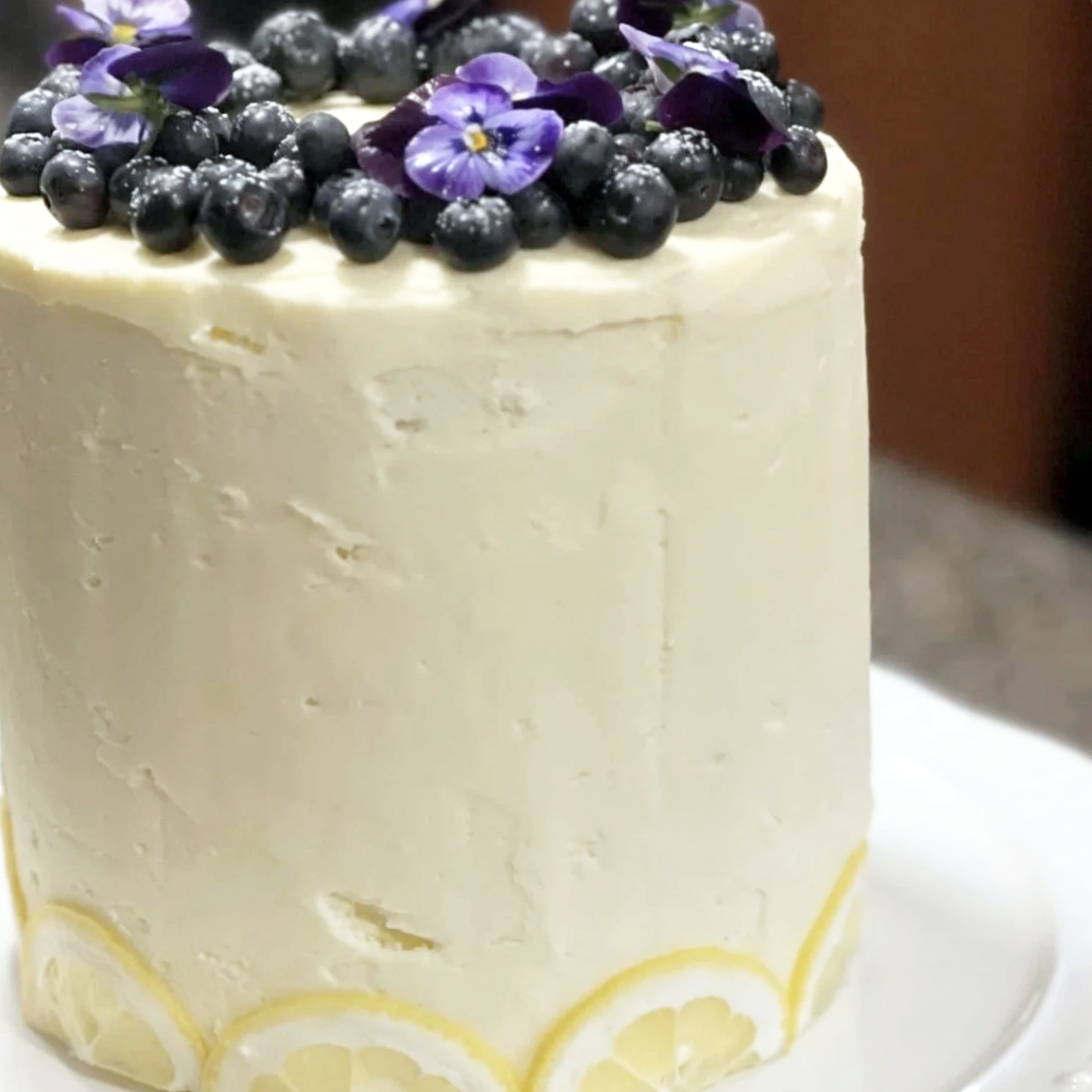 A white frosted cake decorated with blueberries and purple edible flowers on top, with a lemon slice pattern around the base.