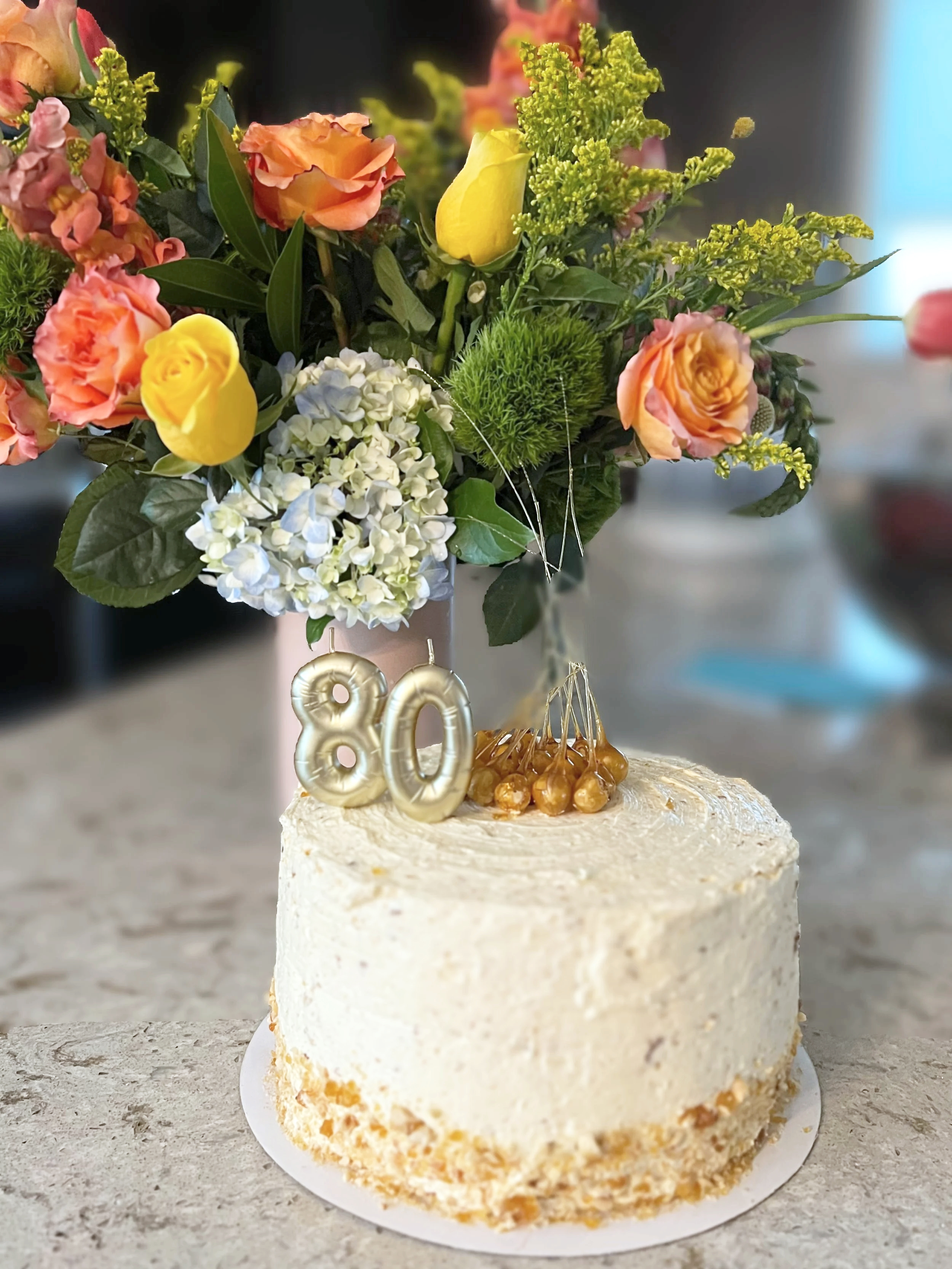 A cake with a white frosting and a crumb coating at the base, decorated with gold '80' candles and caramel-colored candies on top. Behind the cake, there is a bouquet of pink, orange, yellow, and white flowers with green foliage, placed in a pink pot