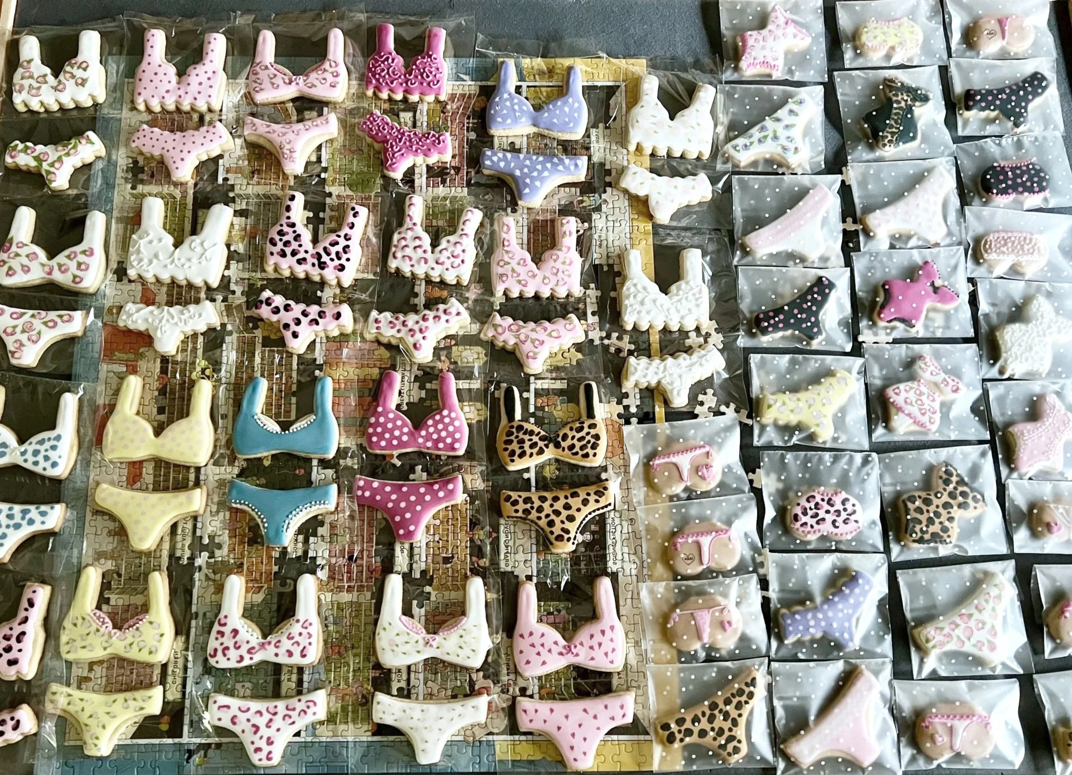 Collection of decorated cookies shaped like bikini tops and bottoms, some in pink, white, yellow, blue, black with colorful polka dots, and leopard print, all arranged on a puzzle with some in clear plastic bags.