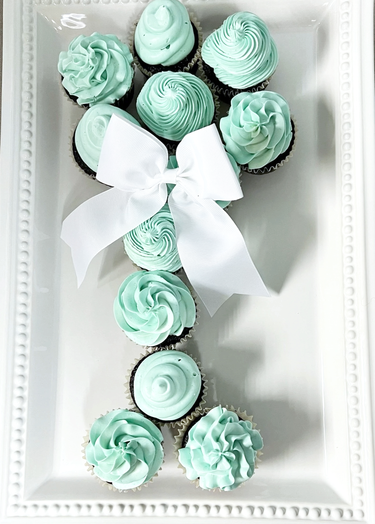 A white rectangular tray with a border of decorative beads, holding cupcakes shaped like the number 1. The cupcakes have mint green frosting with various swirl and rosette designs, and a large white bow decorates the middle.