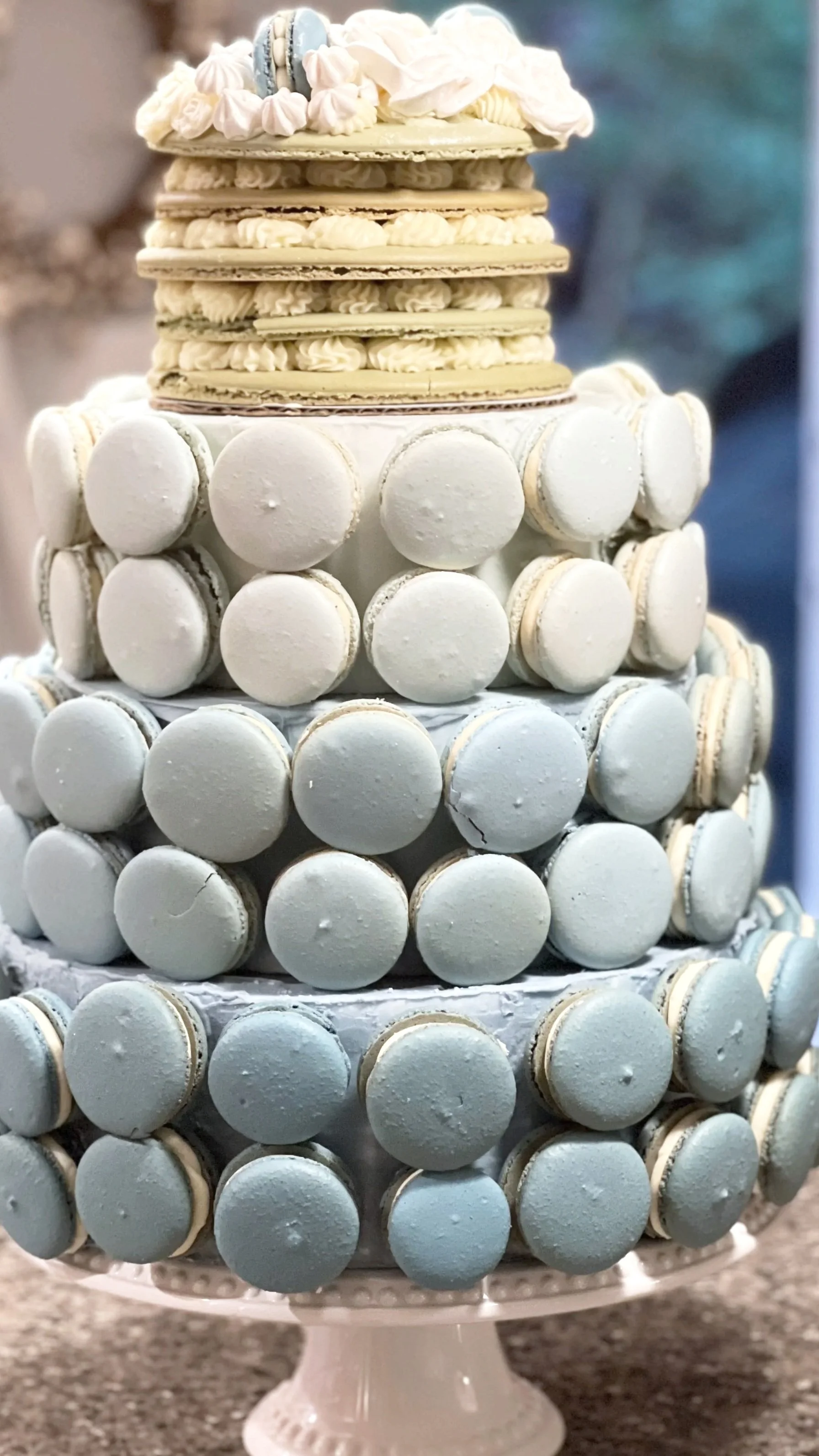 Multi-tiered cake decorated with white macarons and topped with meringue cookies and a blue macaron.
