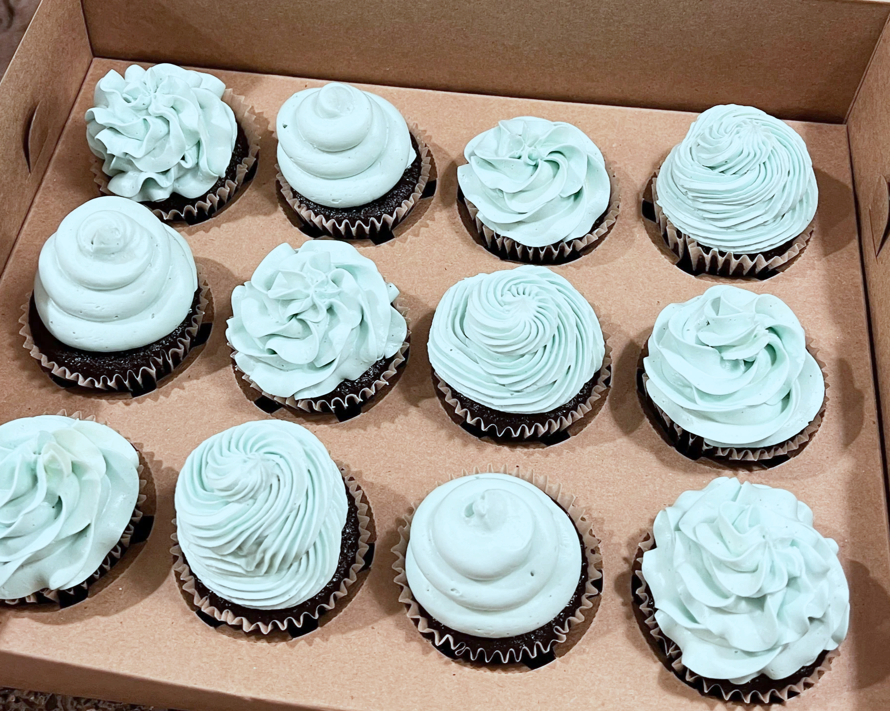 A box of twelve cupcakes decorated with light blue and white frosting in various swirl and rosette designs.