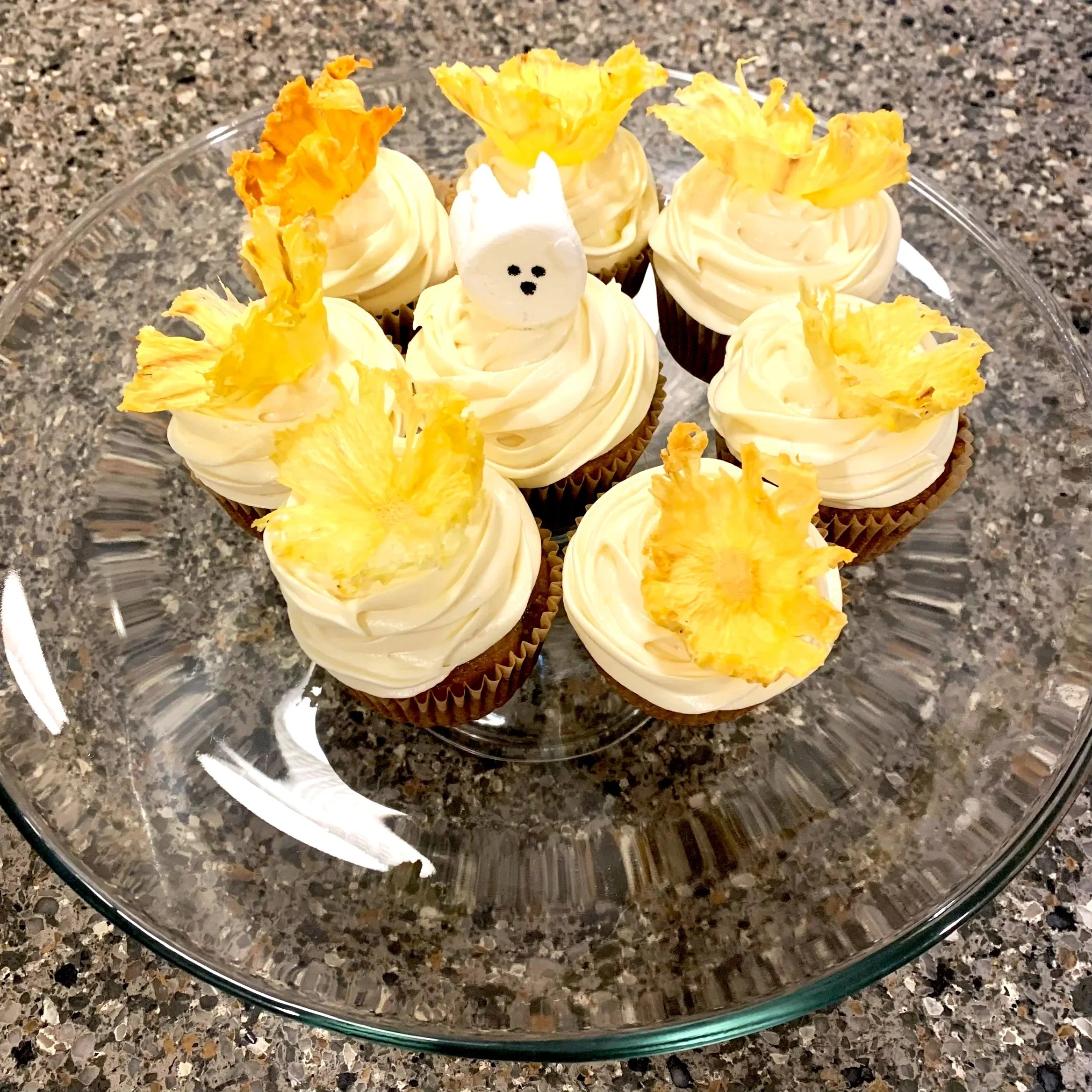 A glass cake stand with ten cupcakes topped with white frosting and pineapple slices as decoration, and one cupcake with a small white ghost face made of marshmallow in the middle.