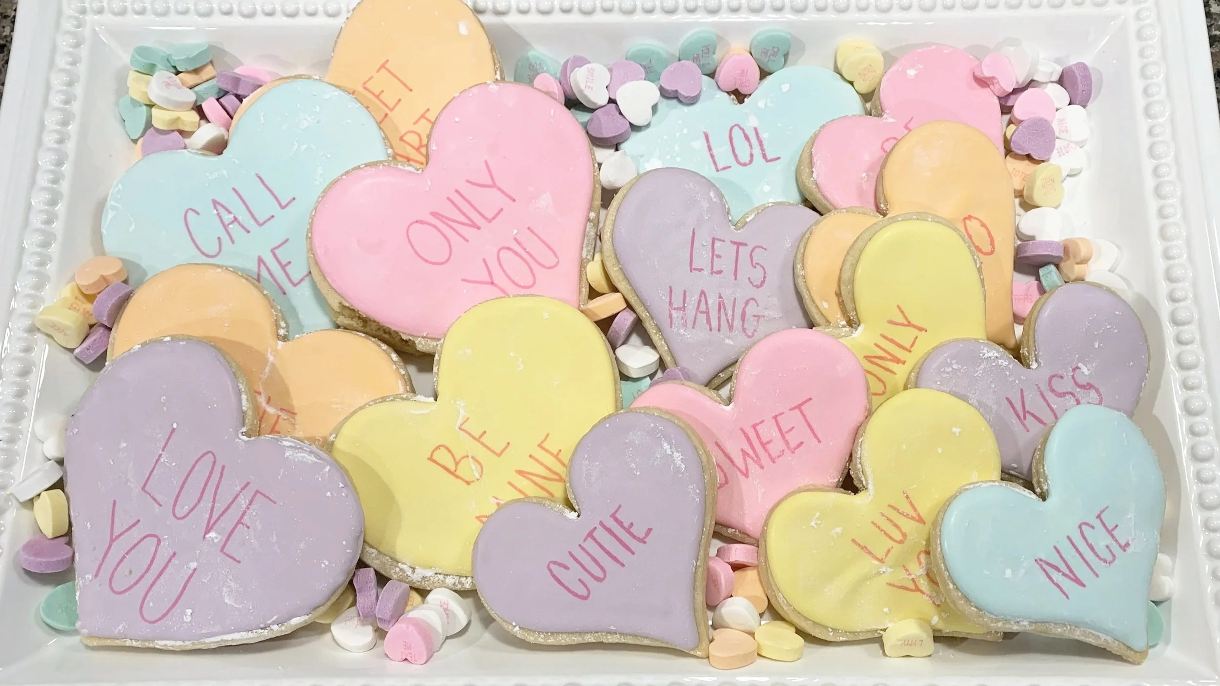 Colorful heart-shaped cookies with various affectionate messages, surrounded by small pastel candies and sprinkles on a white tray.