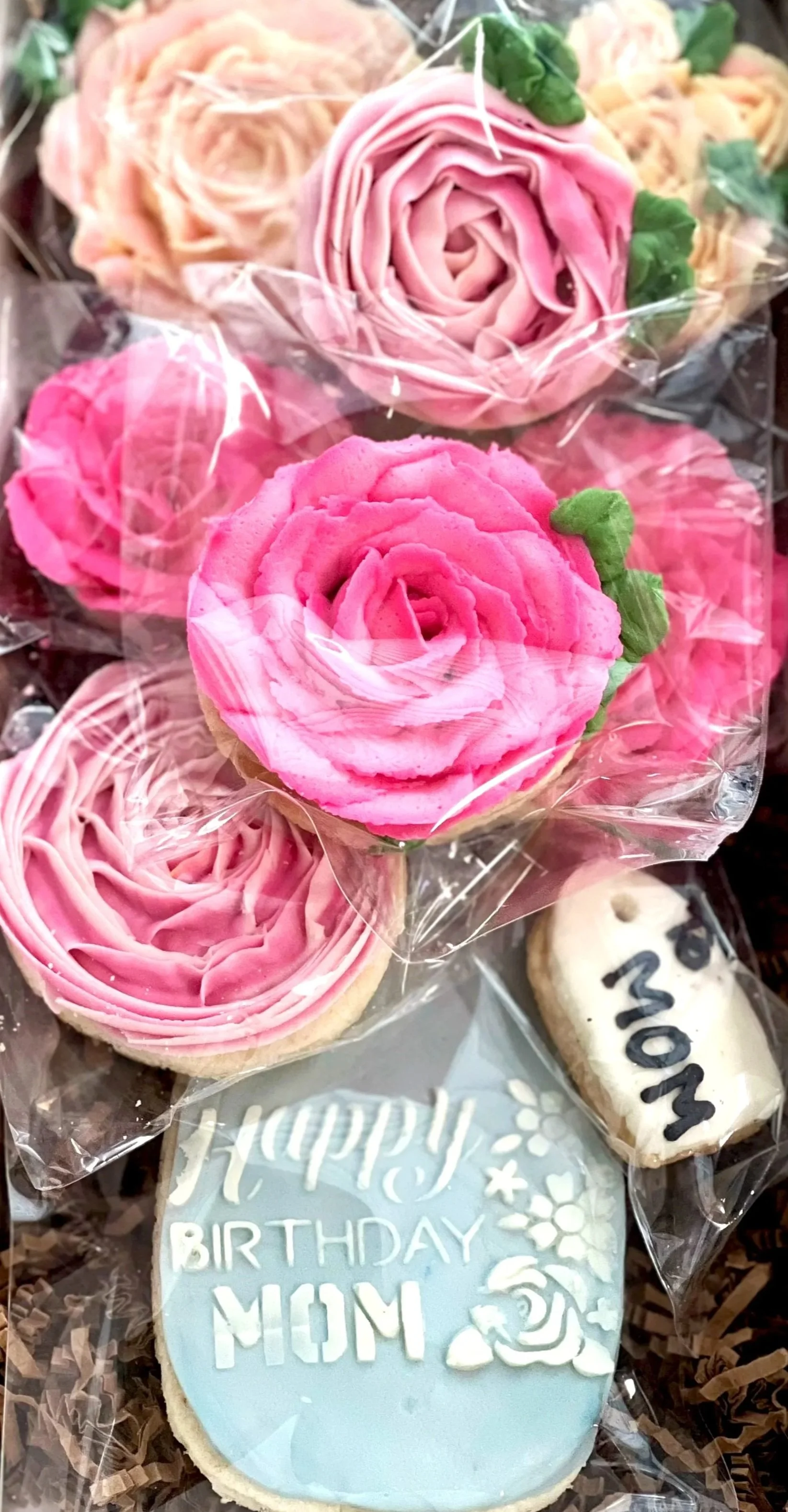 Close-up of decorated cookies and cupcakes wrapped in clear plastic. The cookies include a blue 'Happy Birthday Mom' cookie with white icing and a white flower design, and a rectangular cookie with 'Mom' written in black. The cupcakes are topped with