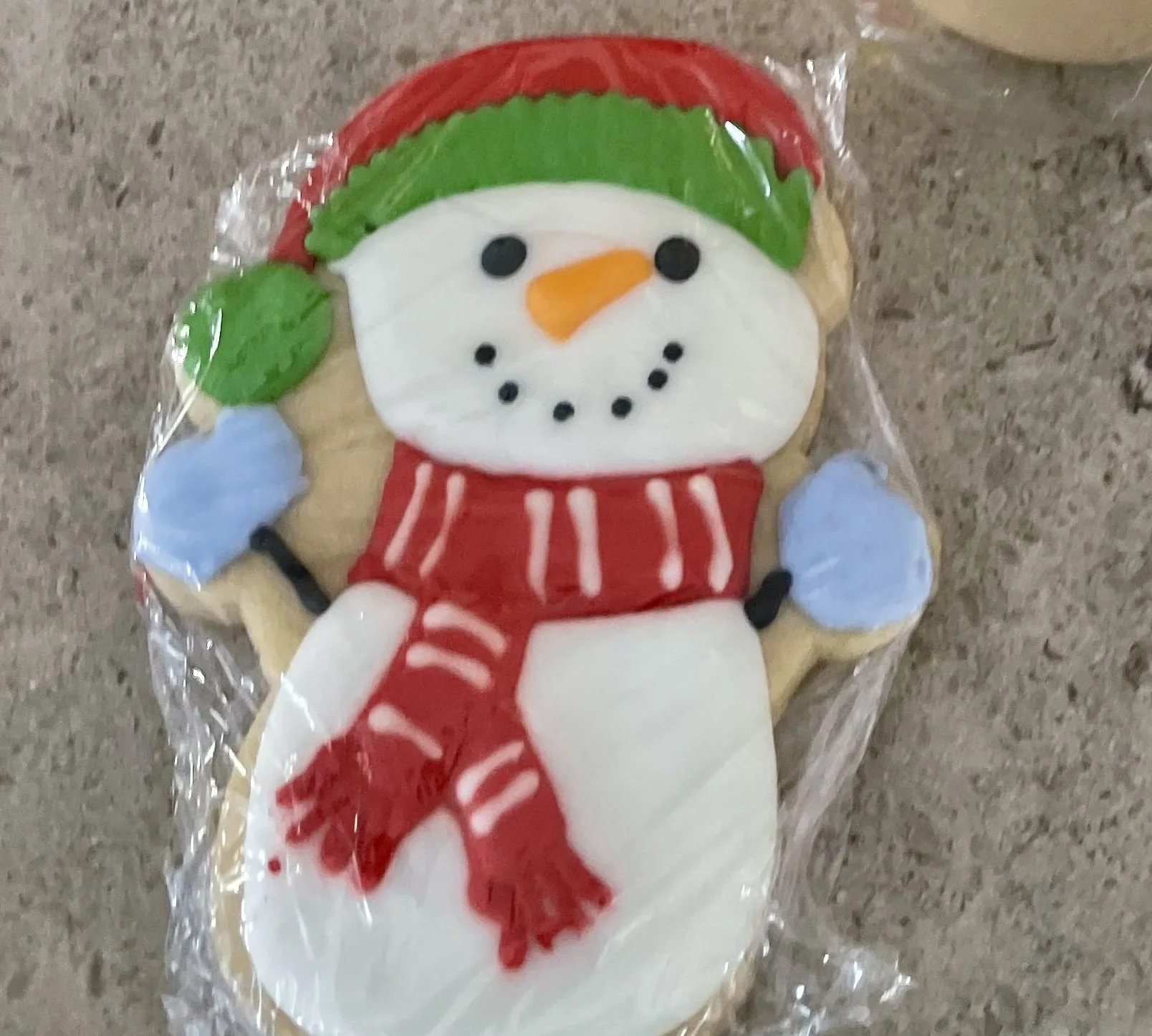 Decorative snowman-shaped cookie with icing, wearing a red scarf and hat, holding blue mittens, wrapped in plastic.