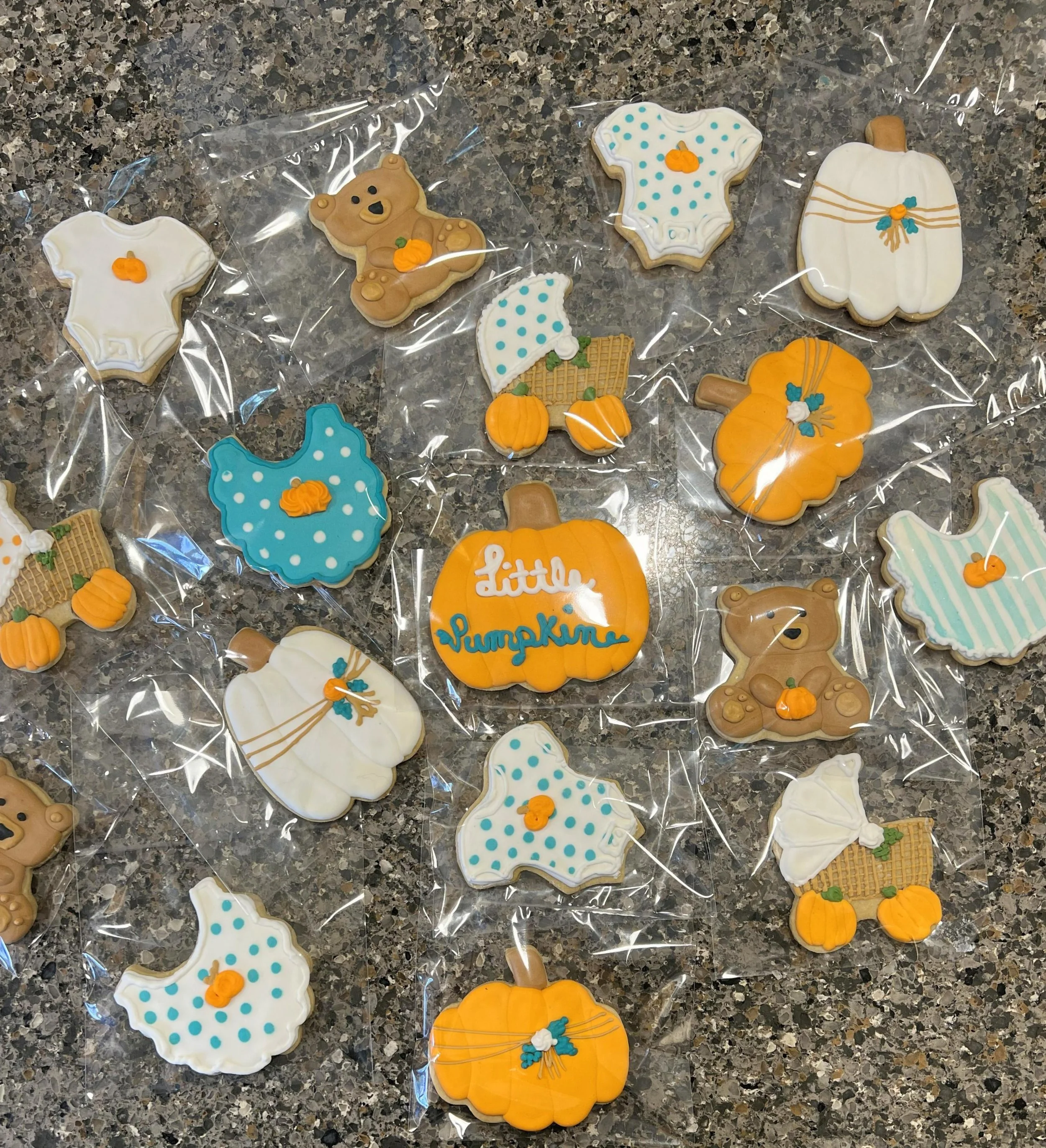 Assorted decorated Halloween cookies on a dark countertop, shaped like pumpkins, teddy bears, baby onesies, umbrellas, and scarecrow pumpkins, with some wrapped in clear plastic.