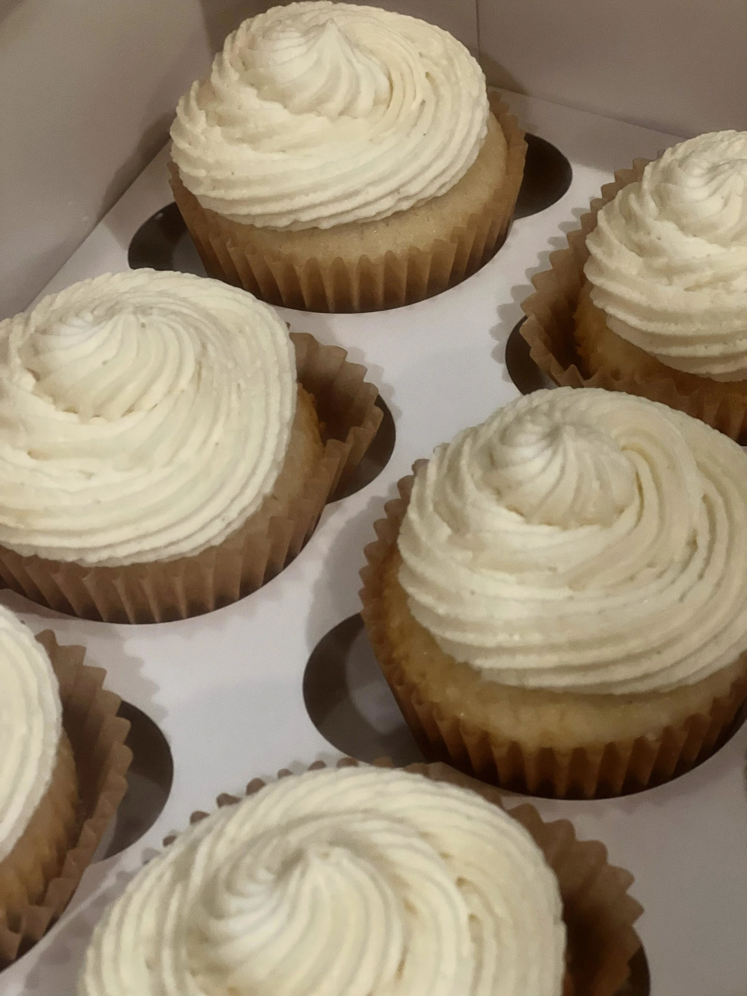 Six vanilla cupcakes with swirl frosting in white cupcake liners inside a box.