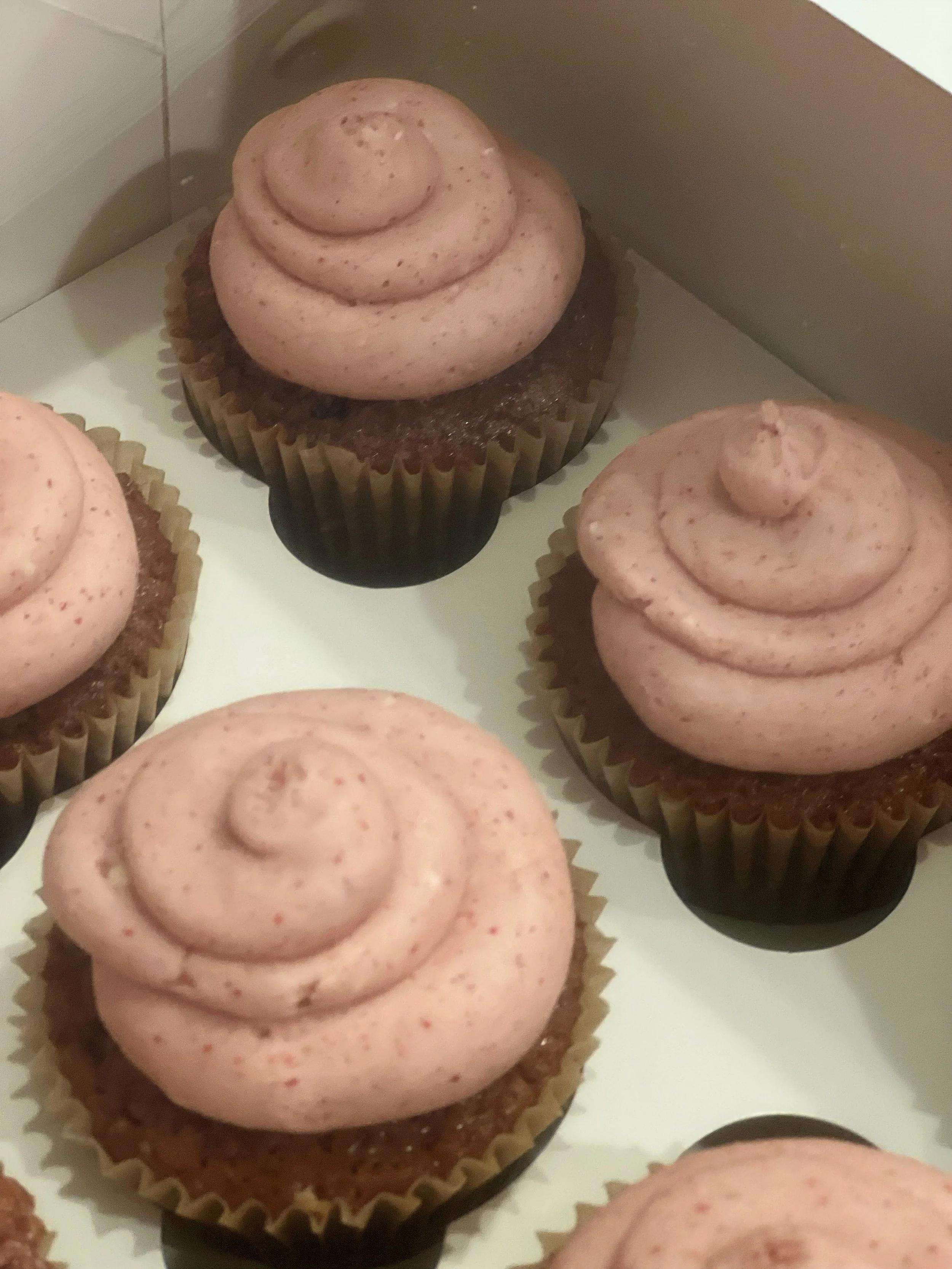Close-up of several cupcakes with pink frosting in paper liners, with some on a tray or box.