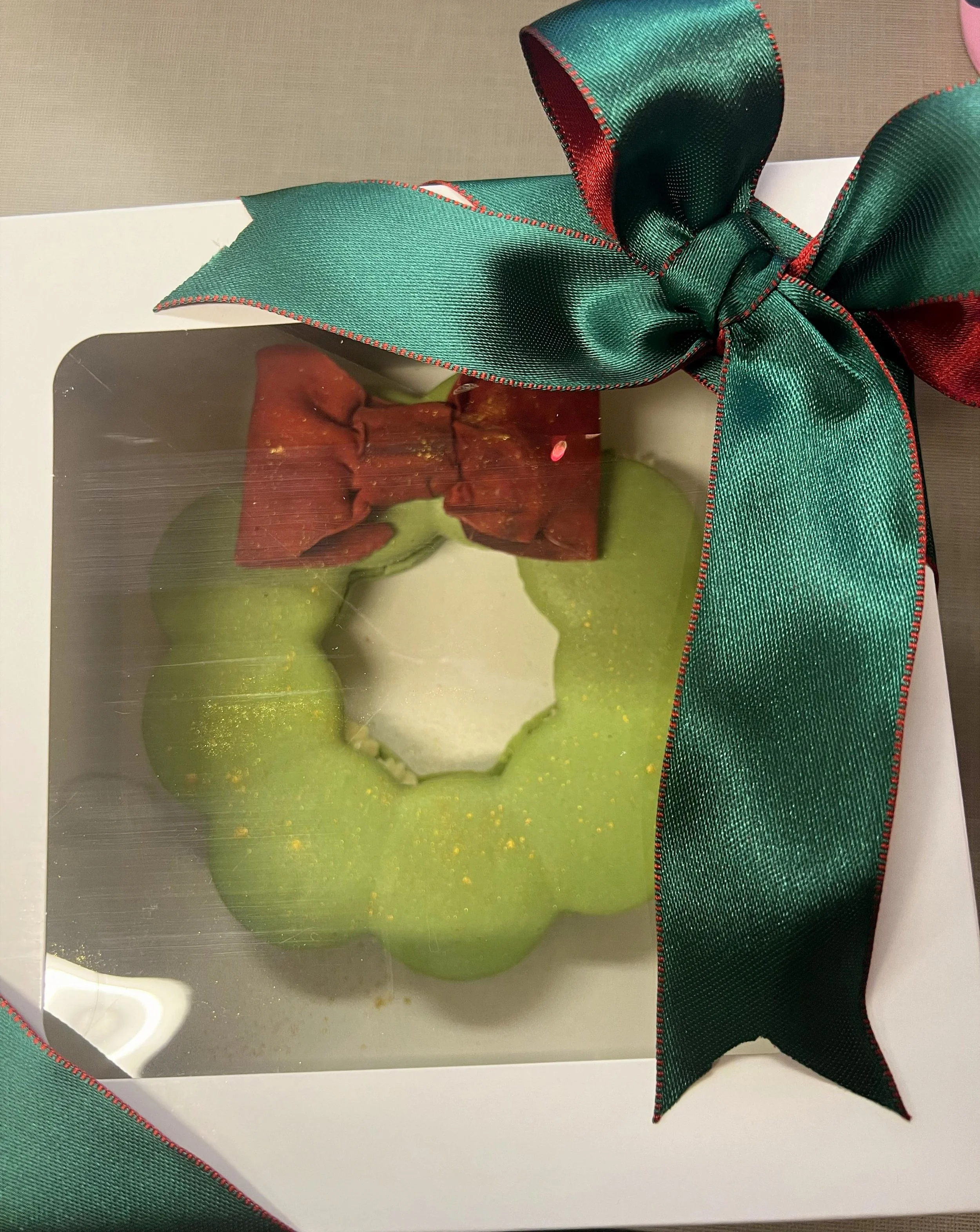 A box containing a green bell-shaped cake decorated with a red ribbon bow, viewed through a clear plastic window.