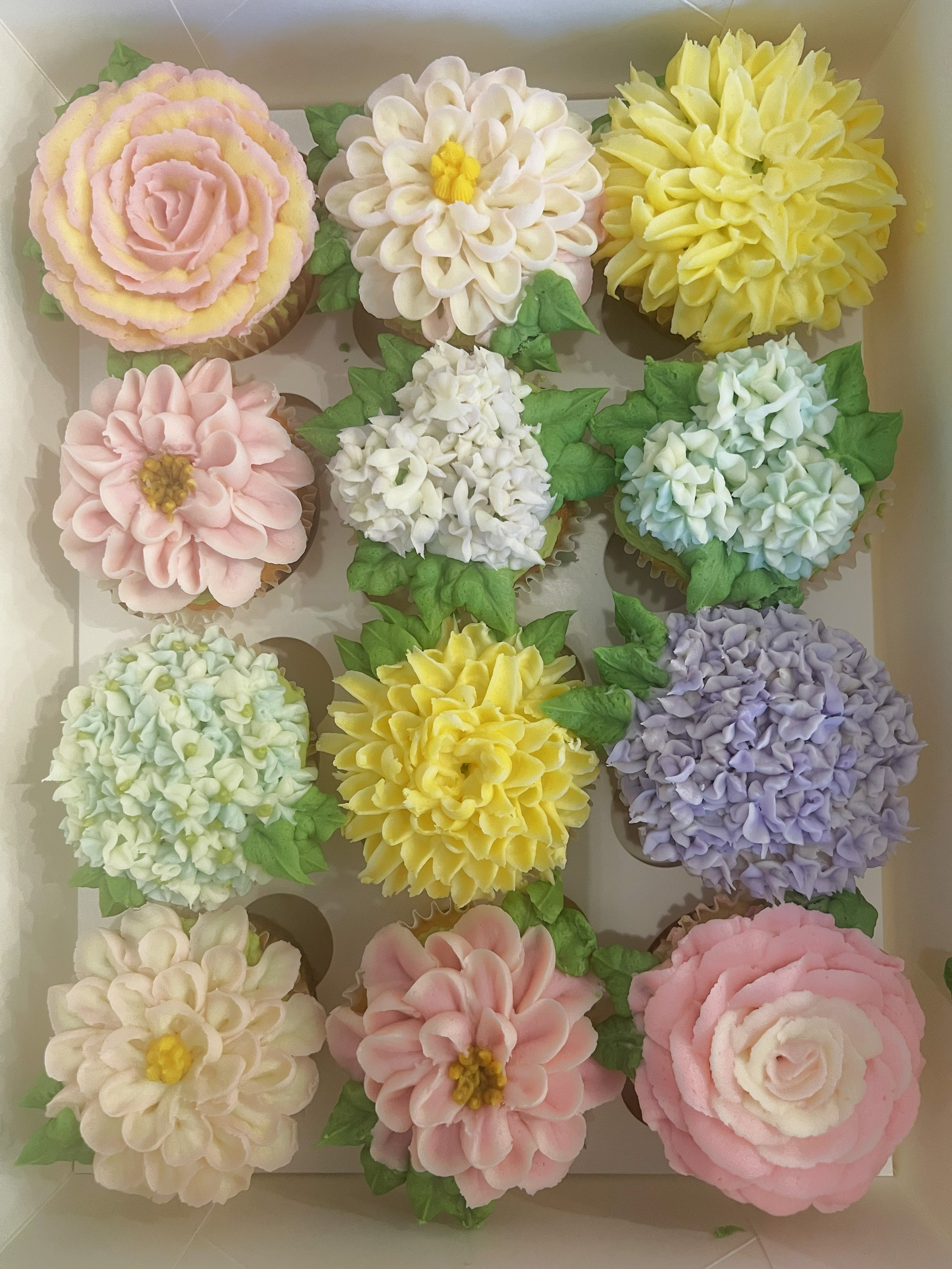 Assorted cupcakes decorated with flower-shaped frosting in pastel colors, including pink, white, yellow, purple, and green, arranged in a grid pattern.
