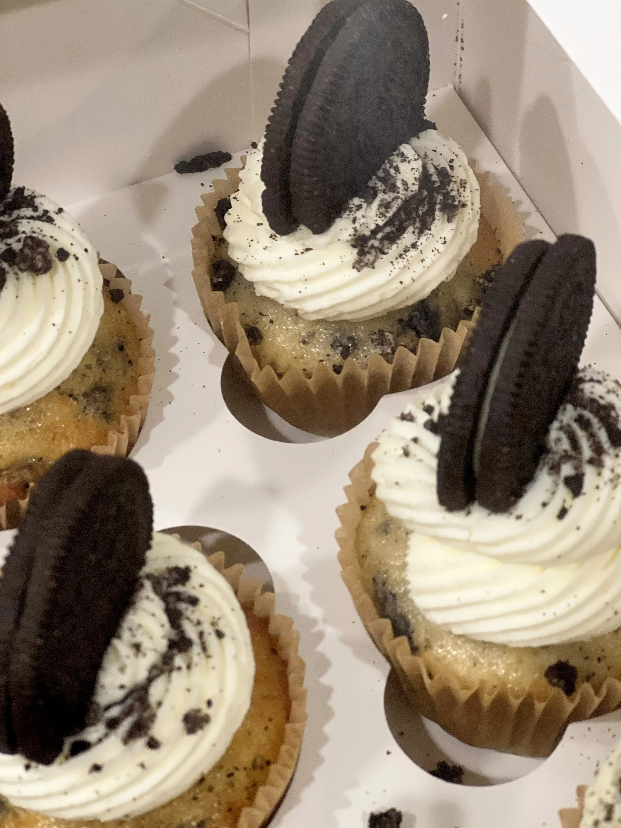 Cupcakes topped with whipped cream, Oreo cookies, and cookie crumbs in a white bakery box.
