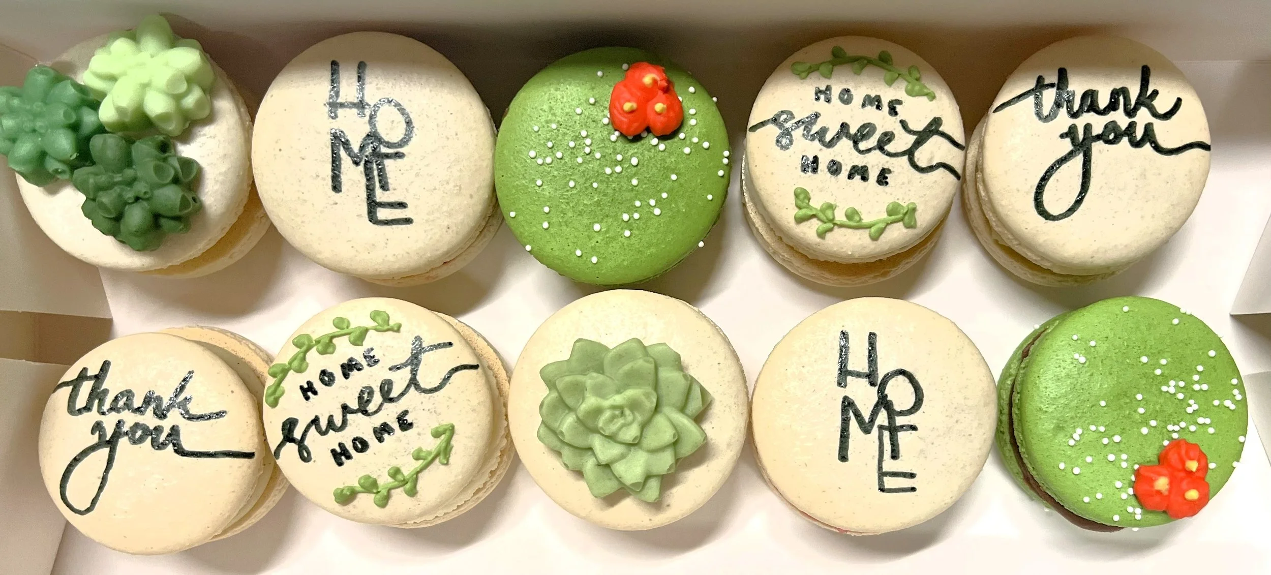 A box of decorated cupcakes with messages and floral designs, including words like "HOME" and "thank you" on white frosted cupcakes, and green frosted cupcakes with floral decorations and small white sprinkles.