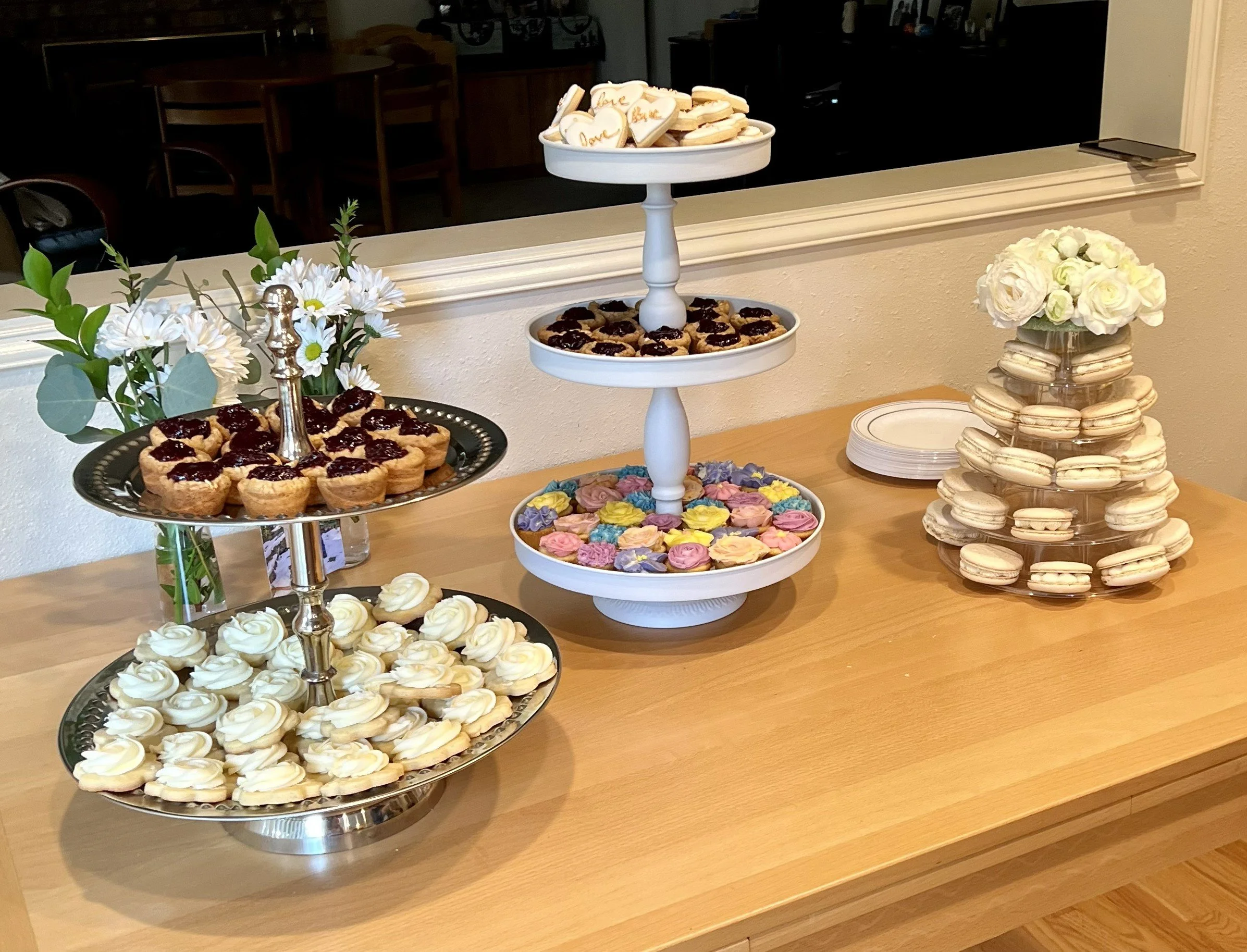 Dessert table with white flower arrangements, macarons, cupcakes, and heart-shaped cookies displayed on silver and white tiered stands.