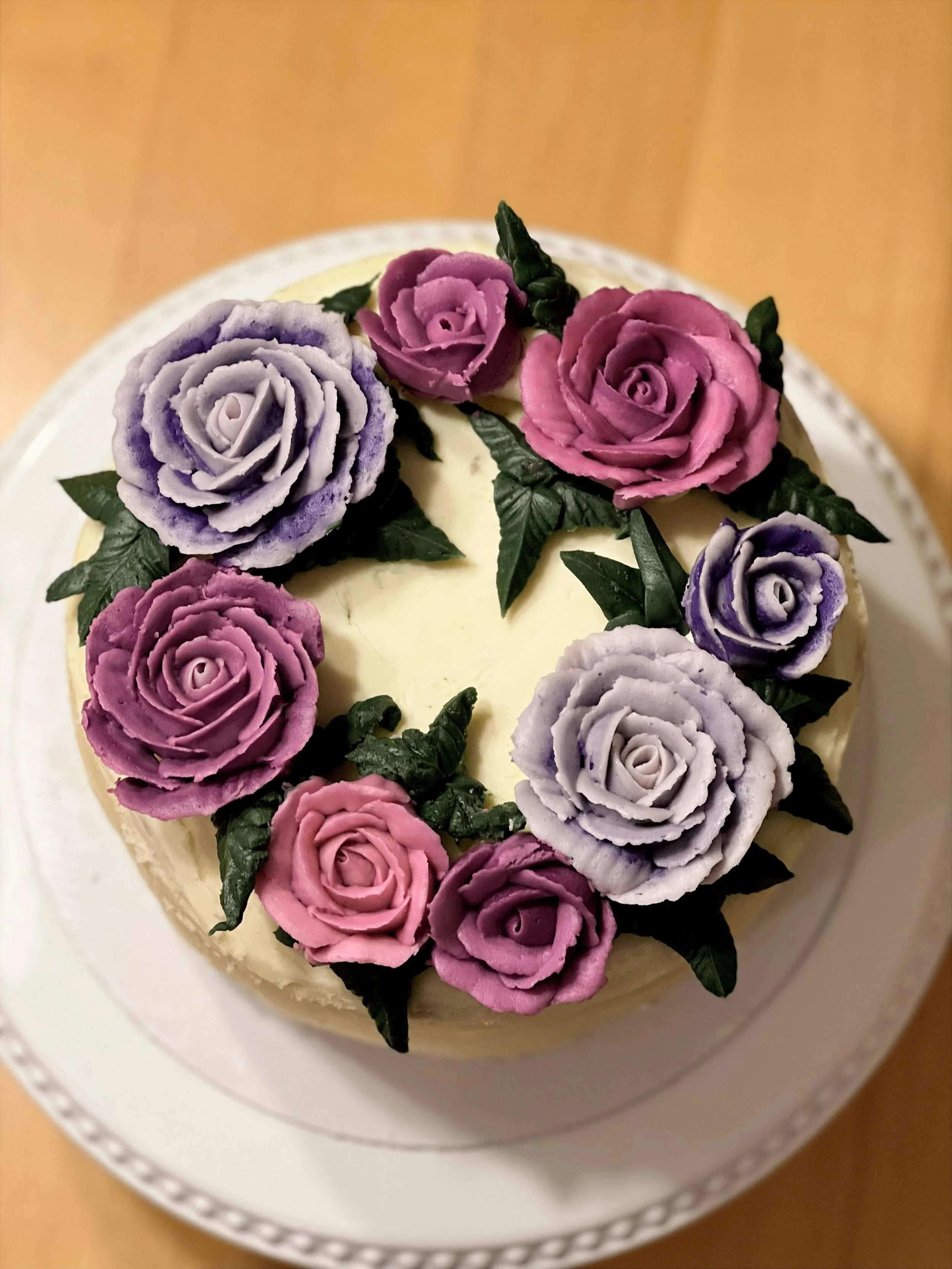 A round cake decorated with pastel-colored roses made of icing, surrounded by green leaves, on a white plate.