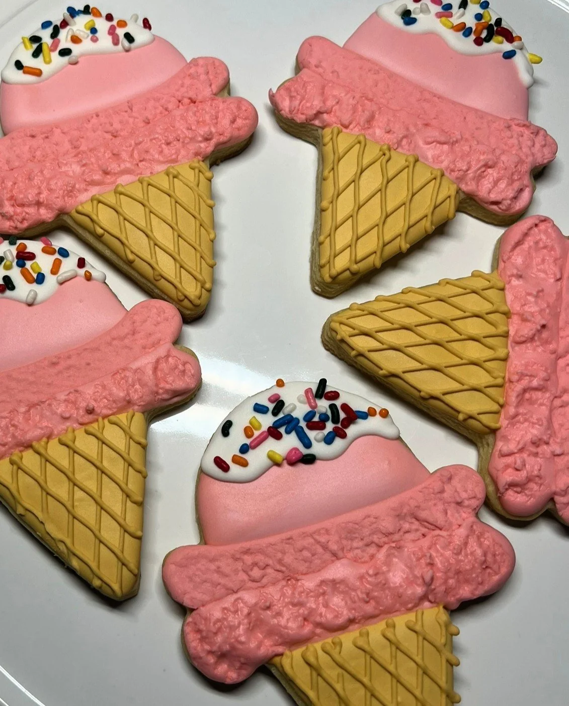 Cookies decorated as ice cream cones with pink and white icing, rainbow sprinkles, and textured pink candy or icing as the ice cream. All arranged on a white plate.