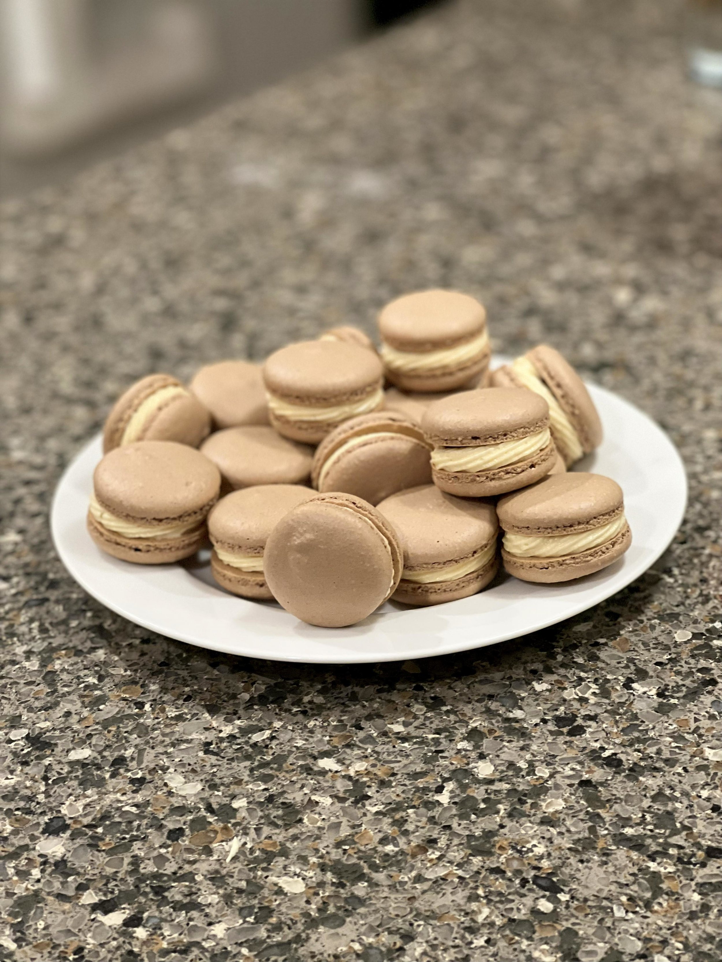 A white plate filled with beige macarons, placed on a speckled granite countertop.