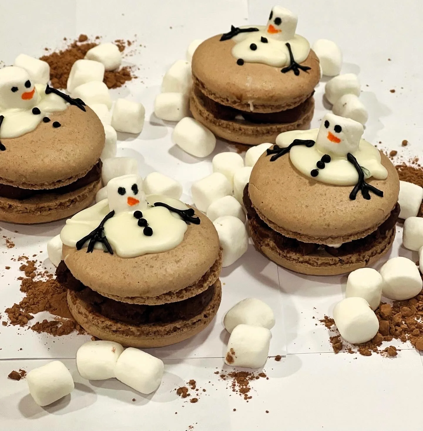 Chocolate macarons decorated with small marshmallows and snowman toppers, surrounded by marshmallows and cocoa powder.