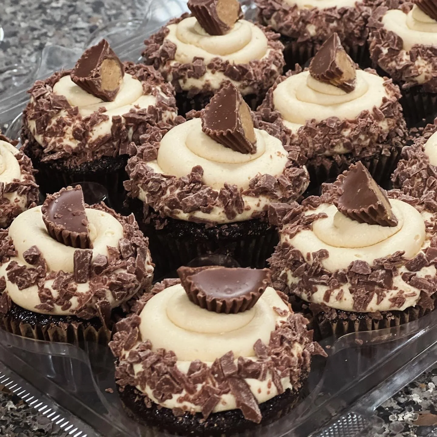 A tray of chocolate cupcakes decorated with vanilla frosting, chopped chocolate pieces, and small chocolate candy halves.