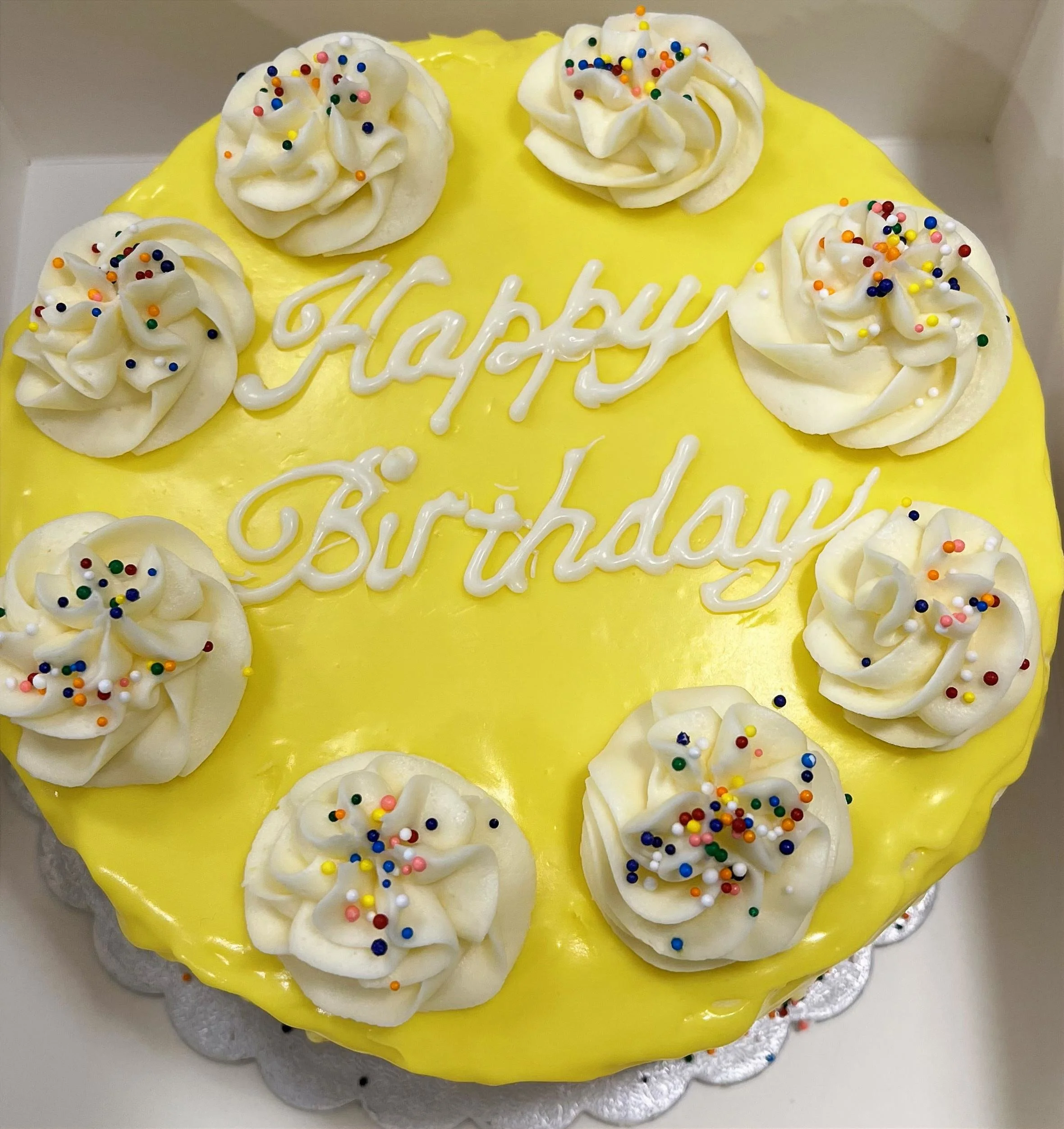 Yellow birthday cake with white frosting swirls topped with colorful sprinkles and the words 'Happy Birthday' written in white icing.