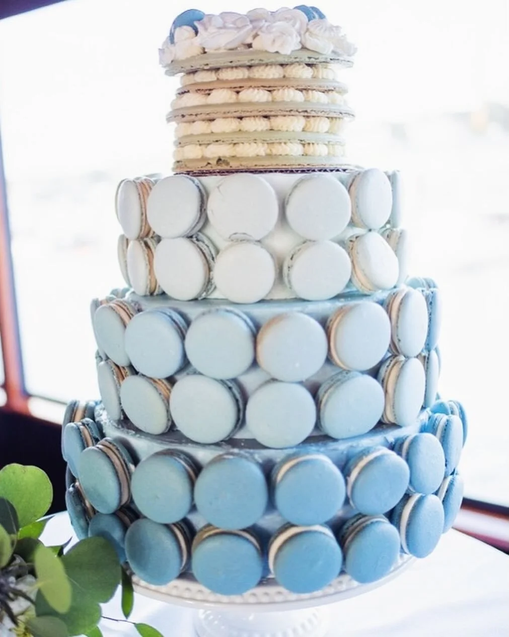 Tiered cake decorated with blue and white macarons and topped with white and blue decorations.