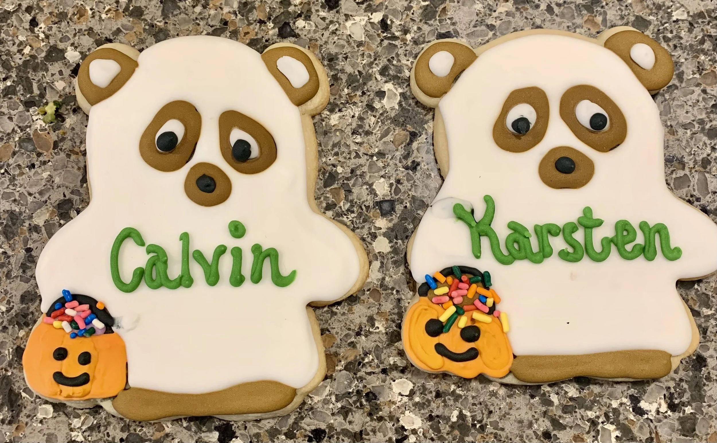 Decorated sugar cookies shaped like bear faces with white icing and brown facial features, each with a small pumpkin decoration and the names 'Calvin' and 'Karsten' written in green icing.