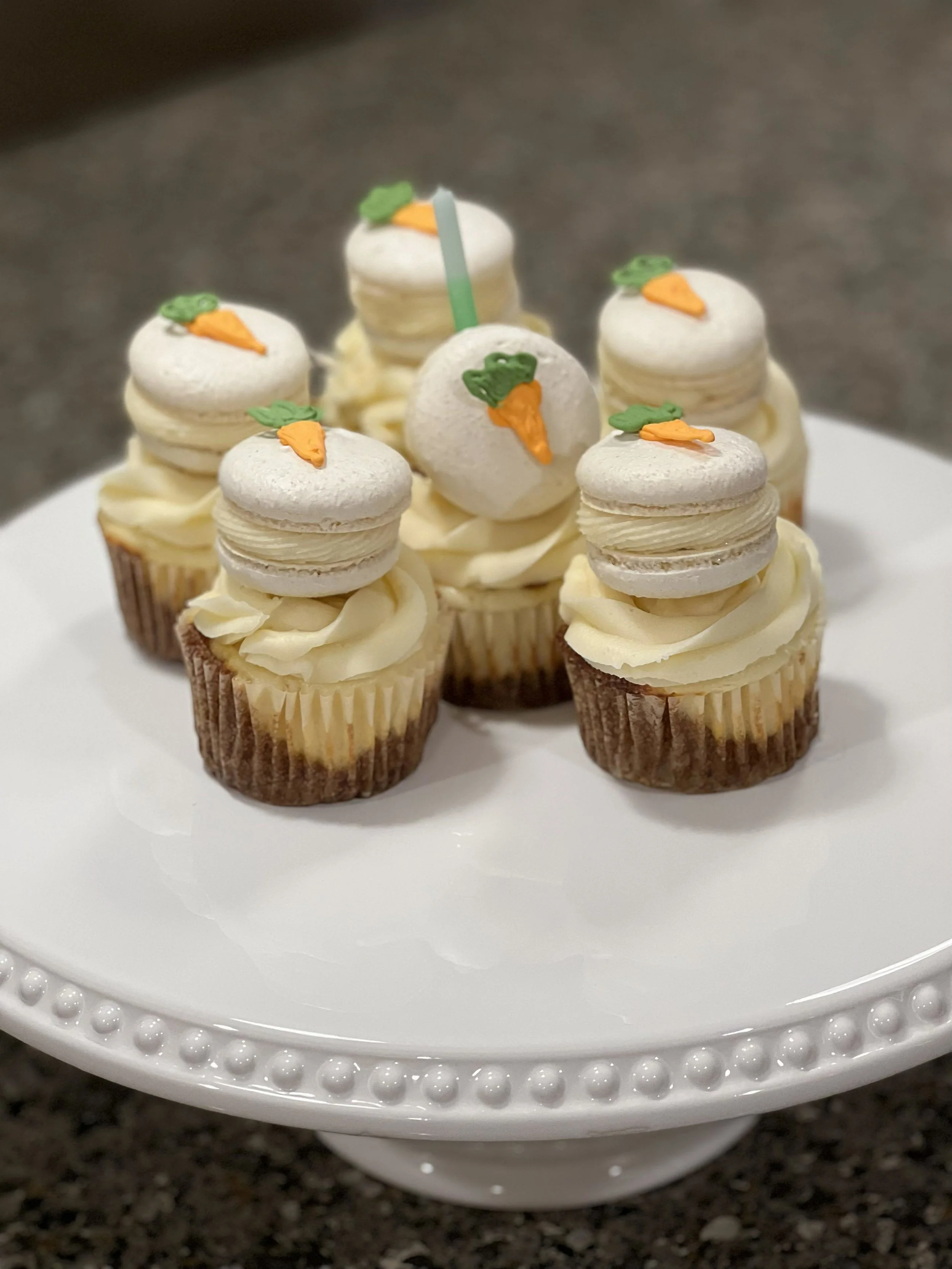 Cupcakes decorated with white frosting, topped with macarons and small carrot decorations, arranged on a white cake stand.