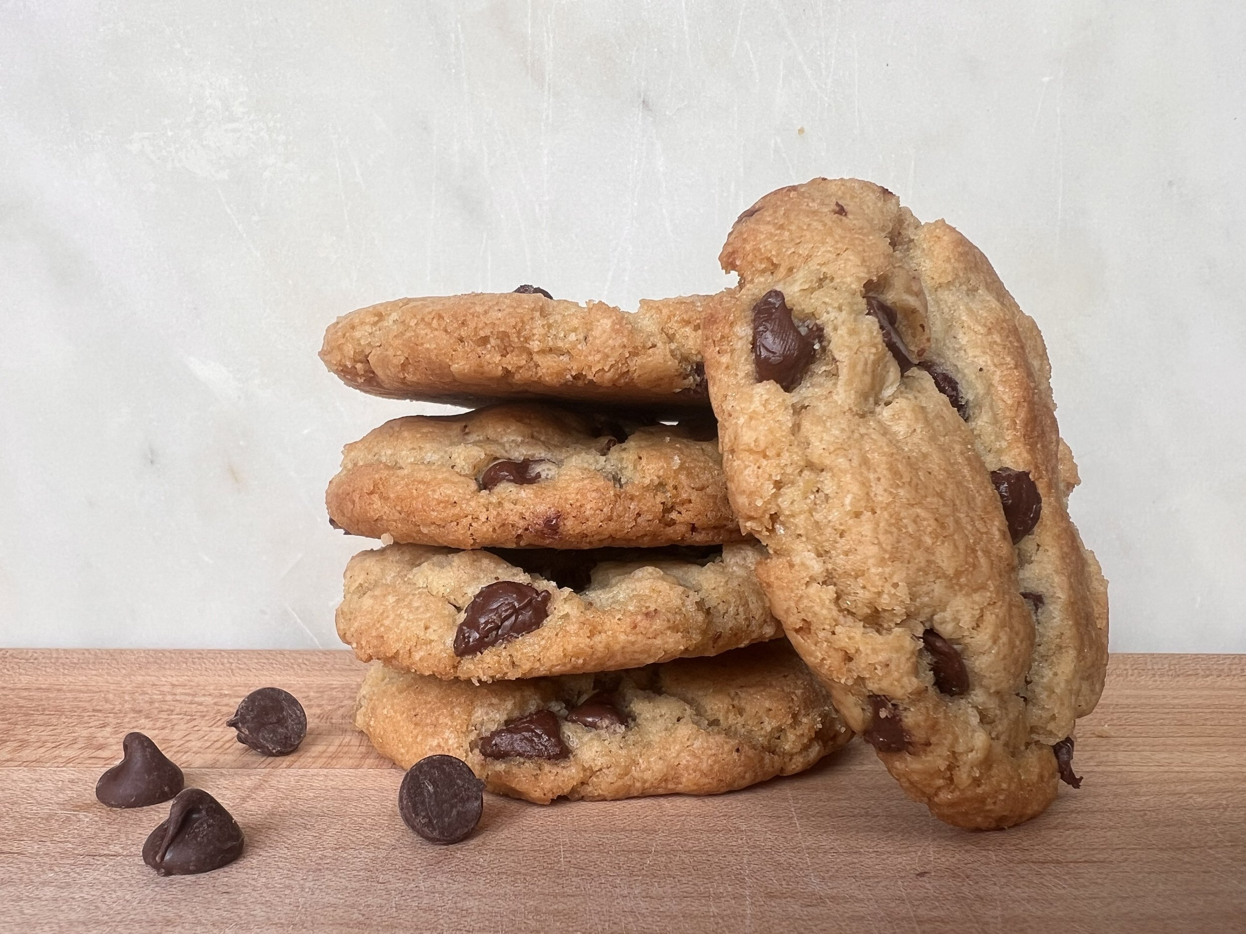 Stack of four chocolate chip cookies on a wooden surface with a few scattered chocolate chips in front, against a plain light background.