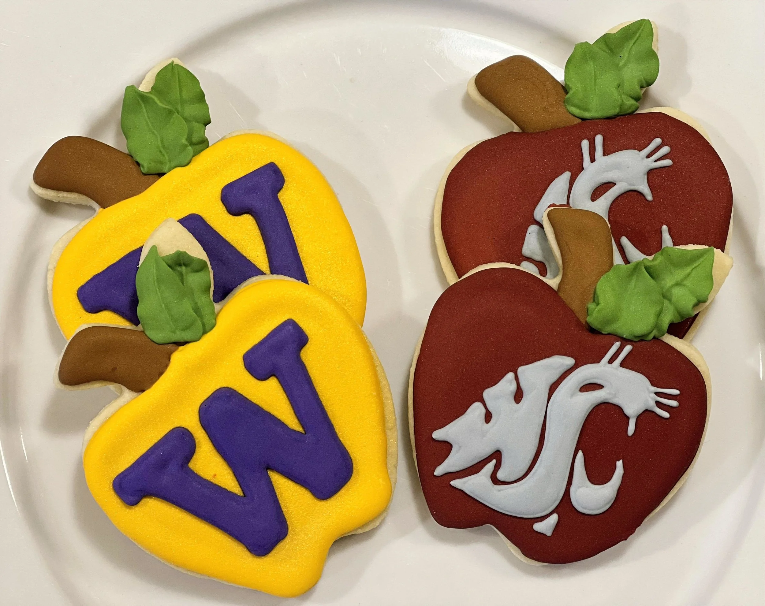 Decorated cookies shaped like the University of Washington husky mascot, with one set featuring the letter W on a yellow background, and the other set with a husky head logo on a red background, each cookie decorated with green leaves and brown paws.