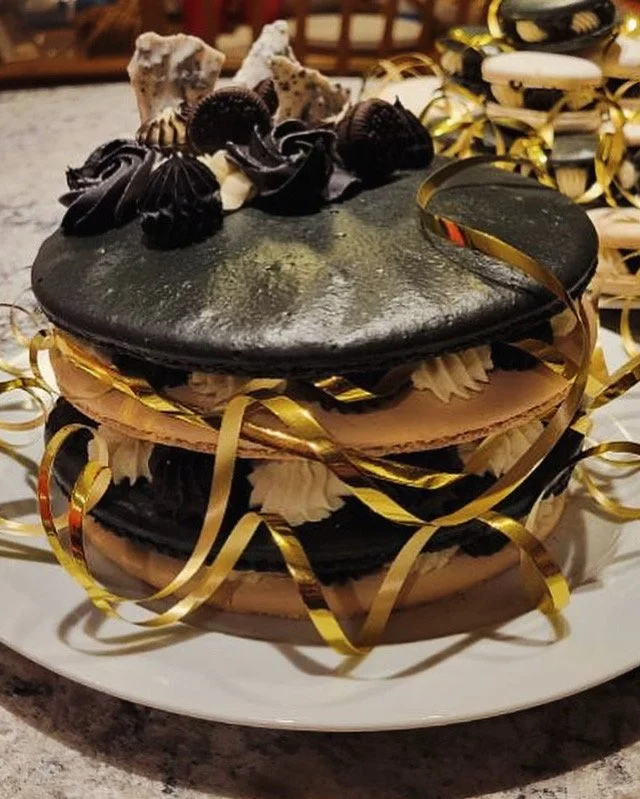 Layered chocolate cake with black, white, and chocolate frosting, topped with decorative chocolate and cookie elements, and surrounded by gold and white ribbon decorations.