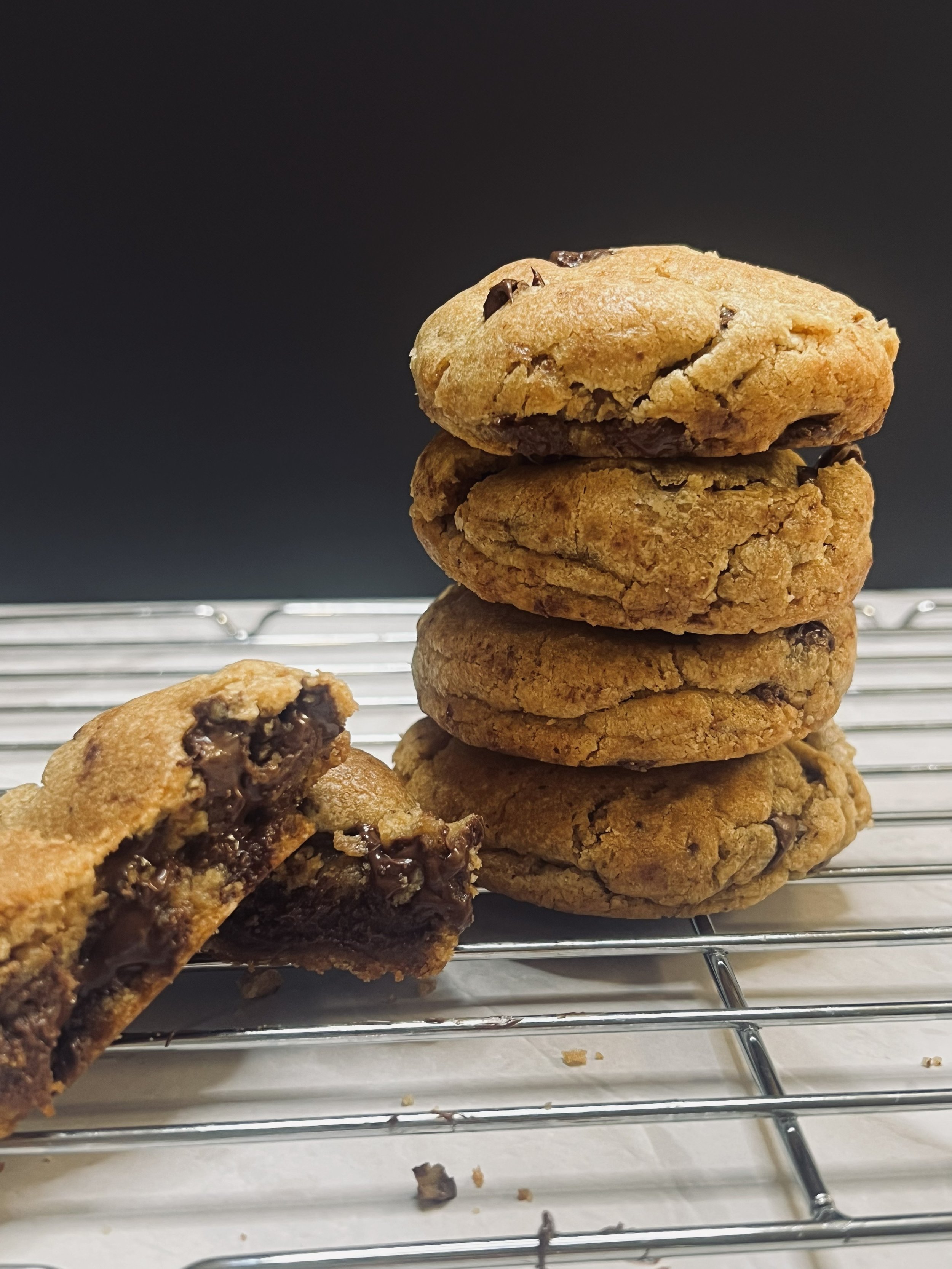 Stack of five chocolate chip cookies on a cooling rack, with one broken cookie in front showing the gooey chocolate inside.