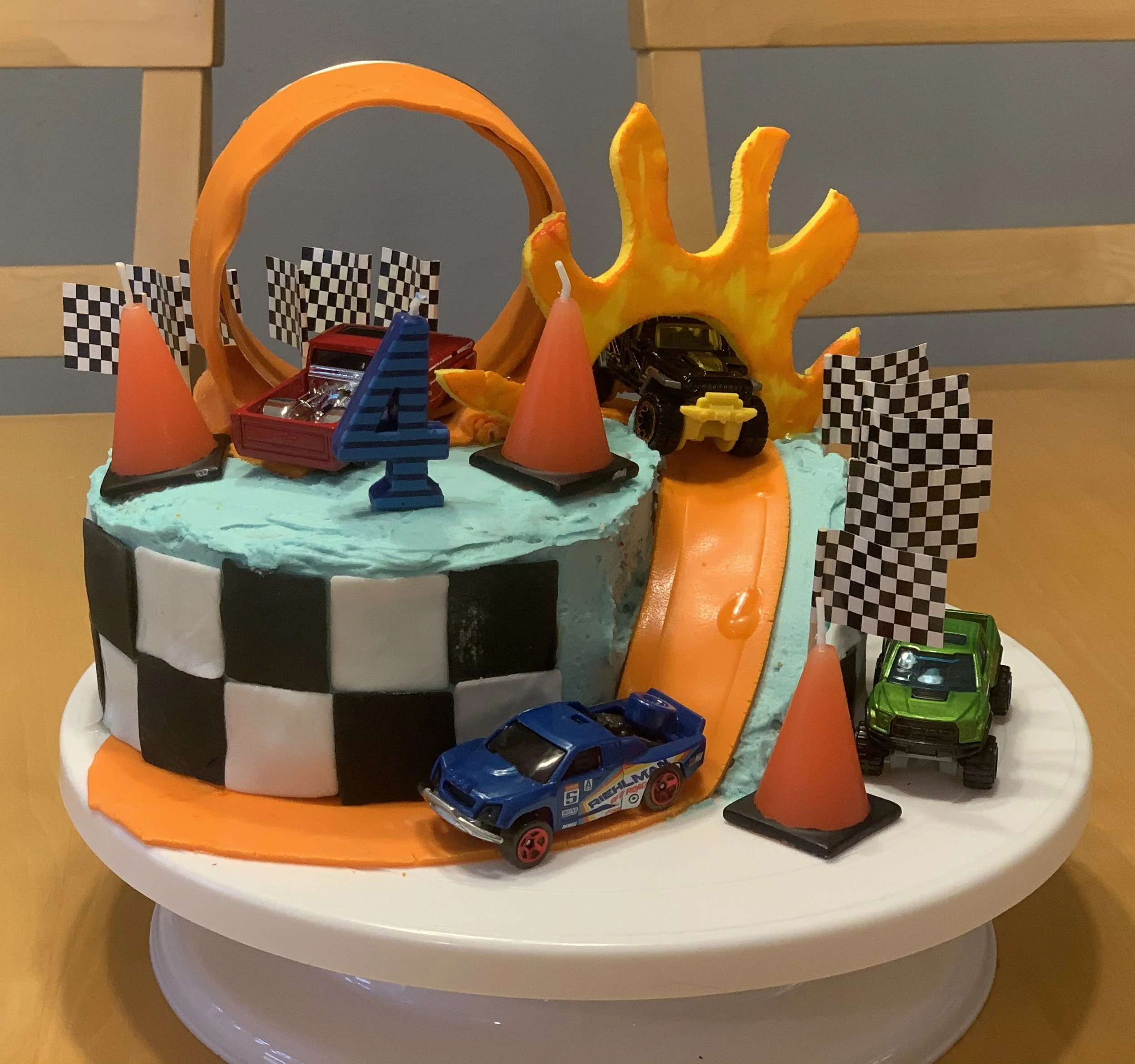 A race car-themed birthday cake decorated with toy race cars, checkered flags, orange cones, and a miniature orange ramp for toy cars, featuring a black and white checkered pattern on the side.