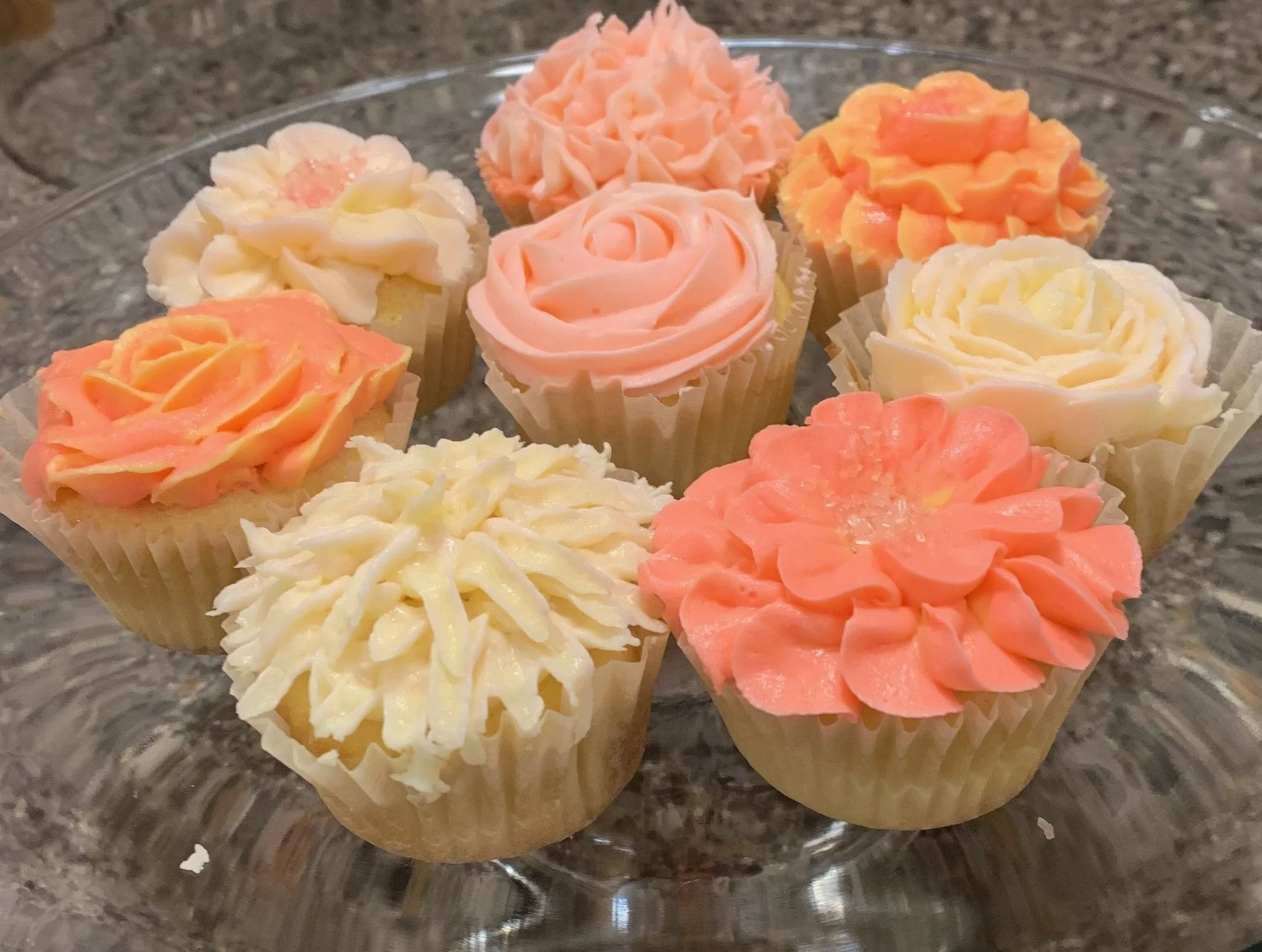 Assorted cupcakes with floral frosting designs on a glass plate, in pink, white, and peach colors.