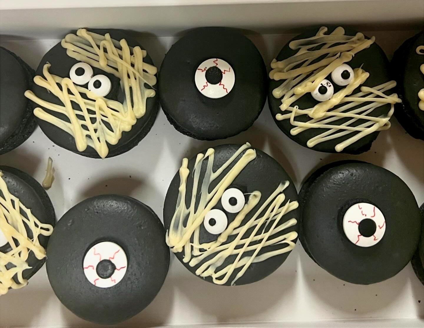 Halloween-themed cupcakes decorated with black icing, white and black edible eyeballs, and white chocolate drizzle resembling zippers and drapes.