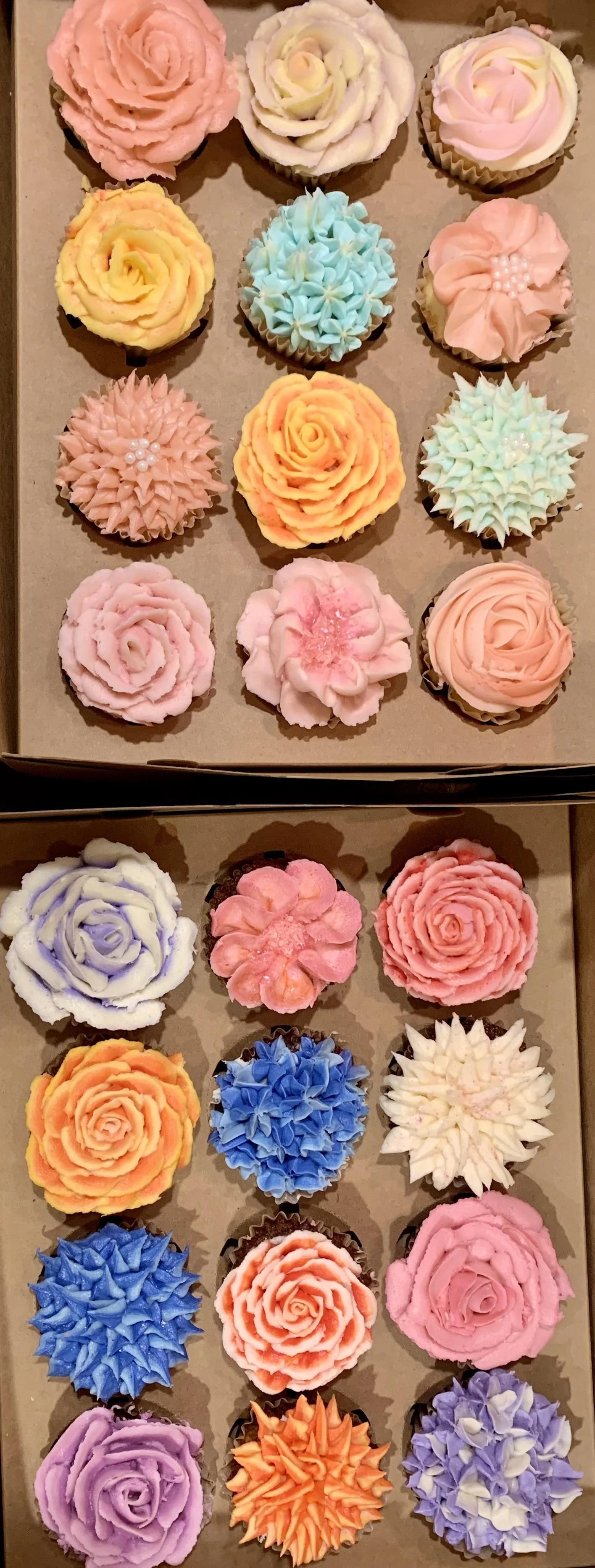 A box of decorated cupcakes with floral and rosette designs in pastel colors, including pink, purple, blue, yellow, and white.