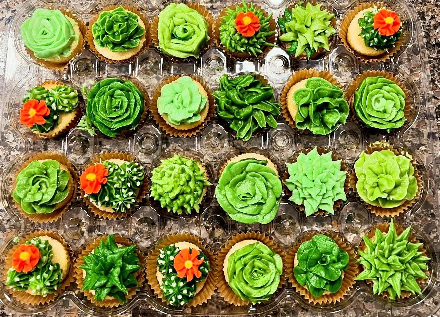 Assorted cupcakes decorated with green frosting in flower and leaf shapes, some with small orange and yellow flowers, arranged in a clear plastic tray on a countertop.