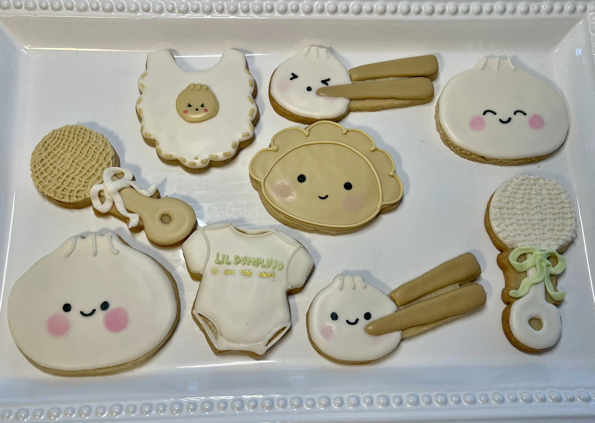 Decorated cookies with cute animal and food designs on a white tray, featuring happy balloon faces, a baby bib, a sheep, a duck, and utensils.