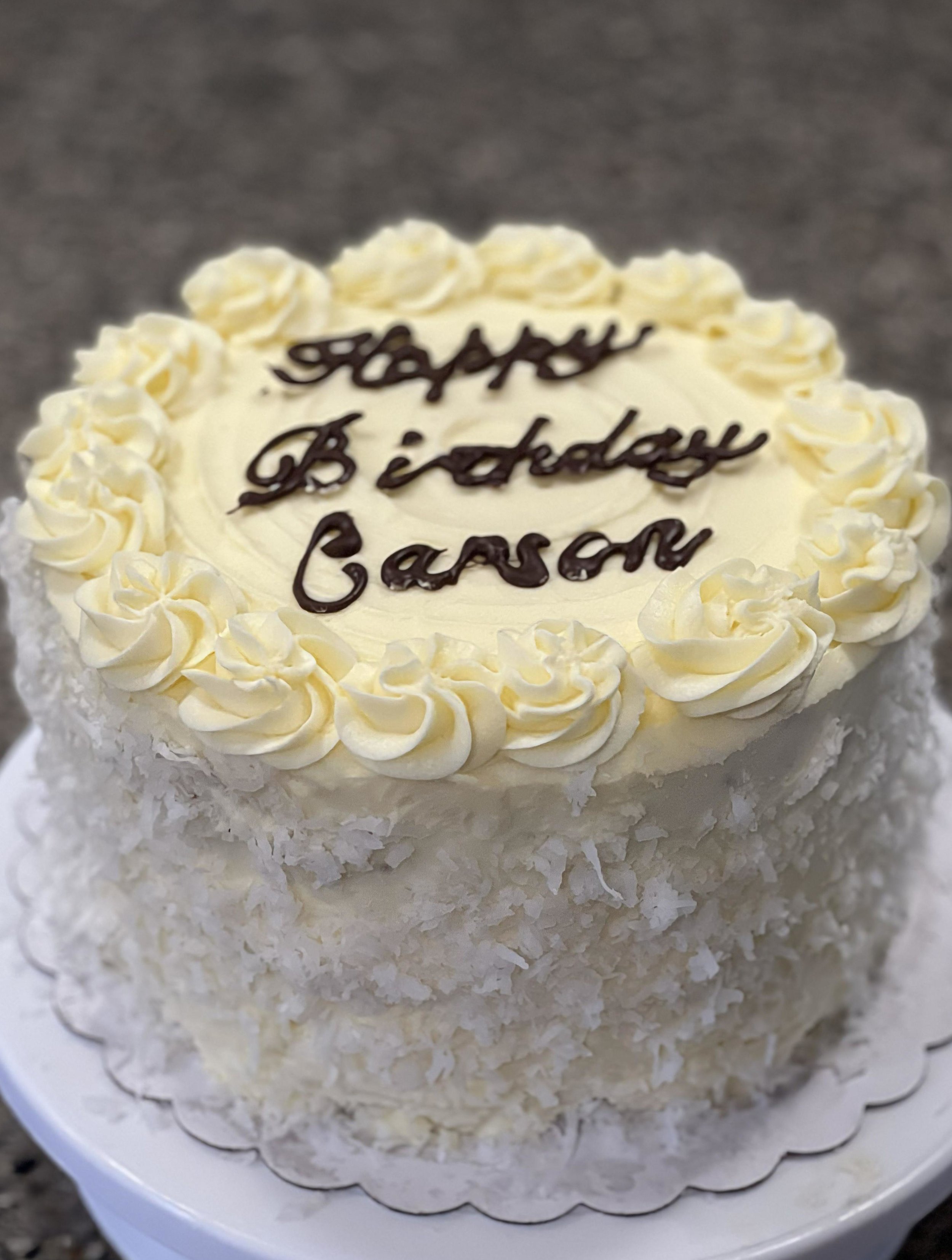 White birthday cake with cream decorations, chocolate writing saying "Happy Birthday Carson" on top, with a textured layer of coconut flakes on the sides, on a white cake stand.
